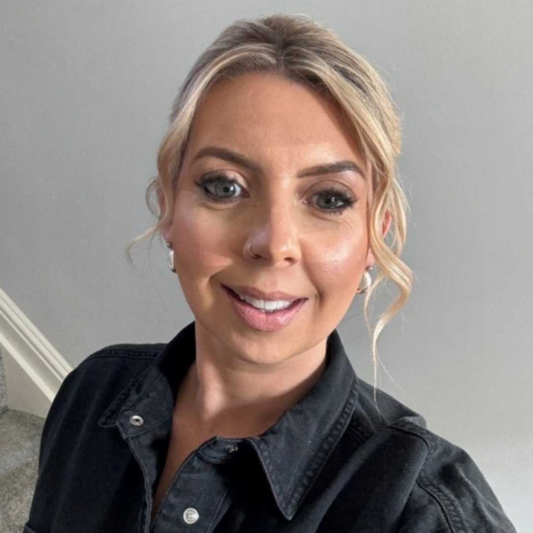 Smiling person in a dark jacket taking a selfie indoors against a light wall