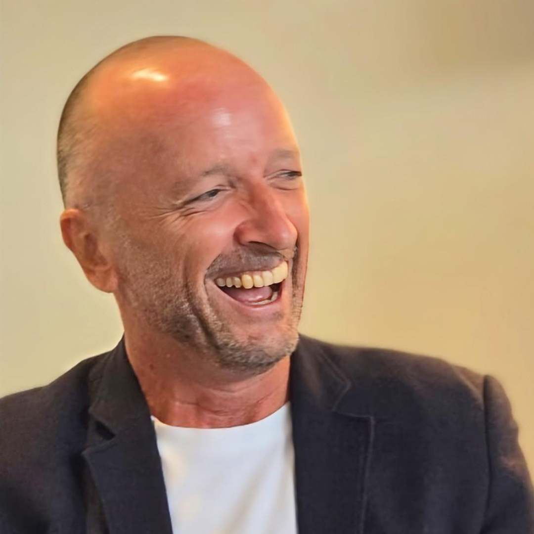 Smiling bald man in a dark blazer and white shirt, laughing against a warm indoor background