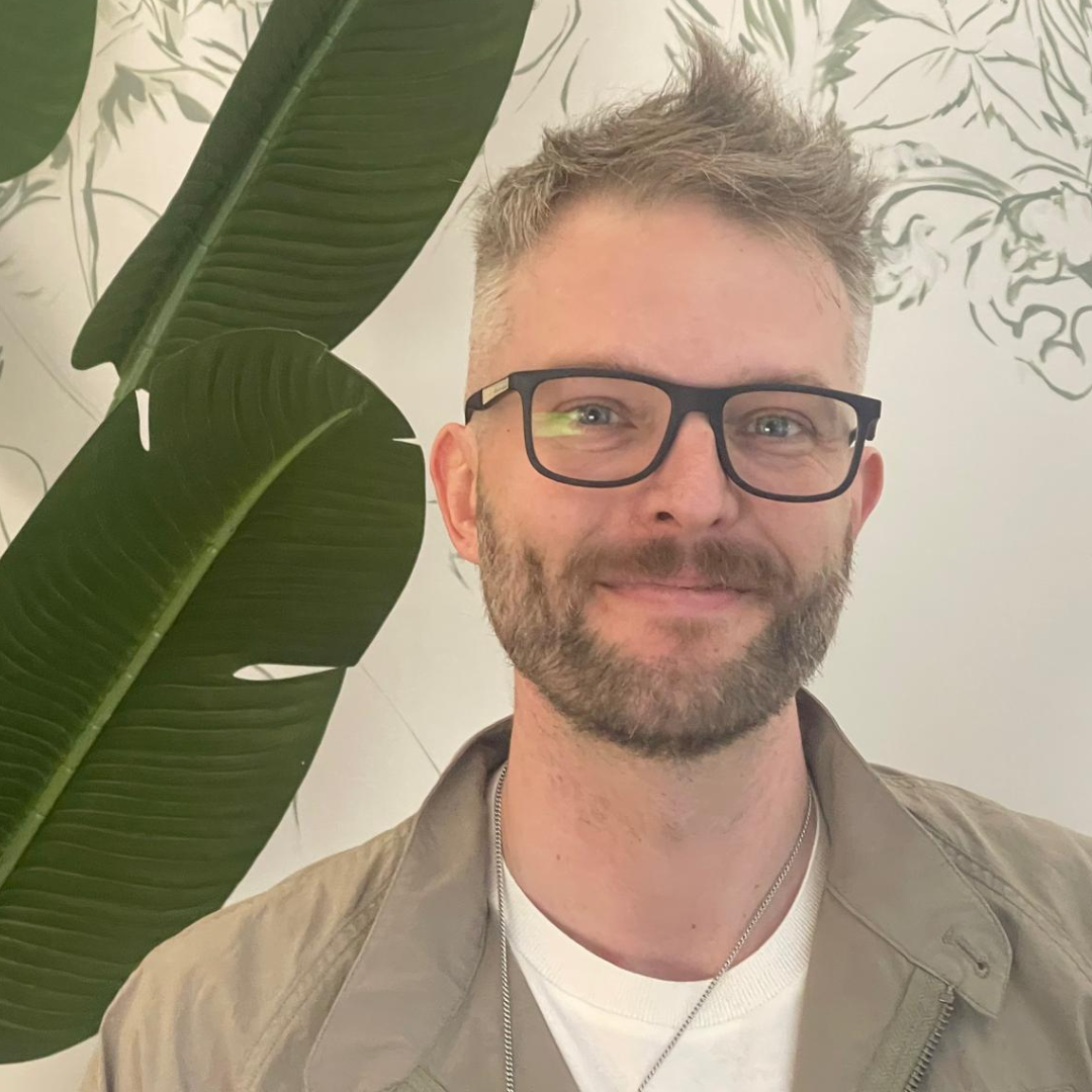 Smiling person with glasses and short beard, standing indoors beside a large green leaf plant.