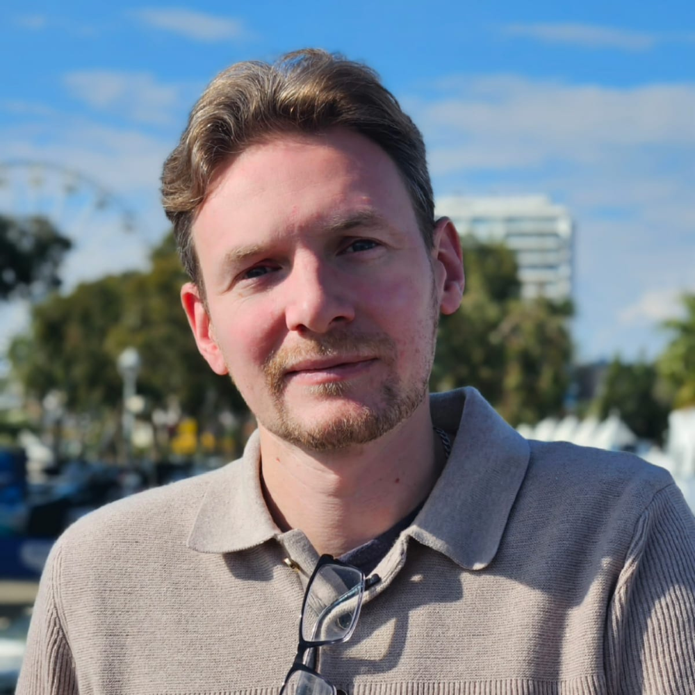 Portrait of a man outdoors under a blue sky, wearing a beige sweater and glasses around his neck.
