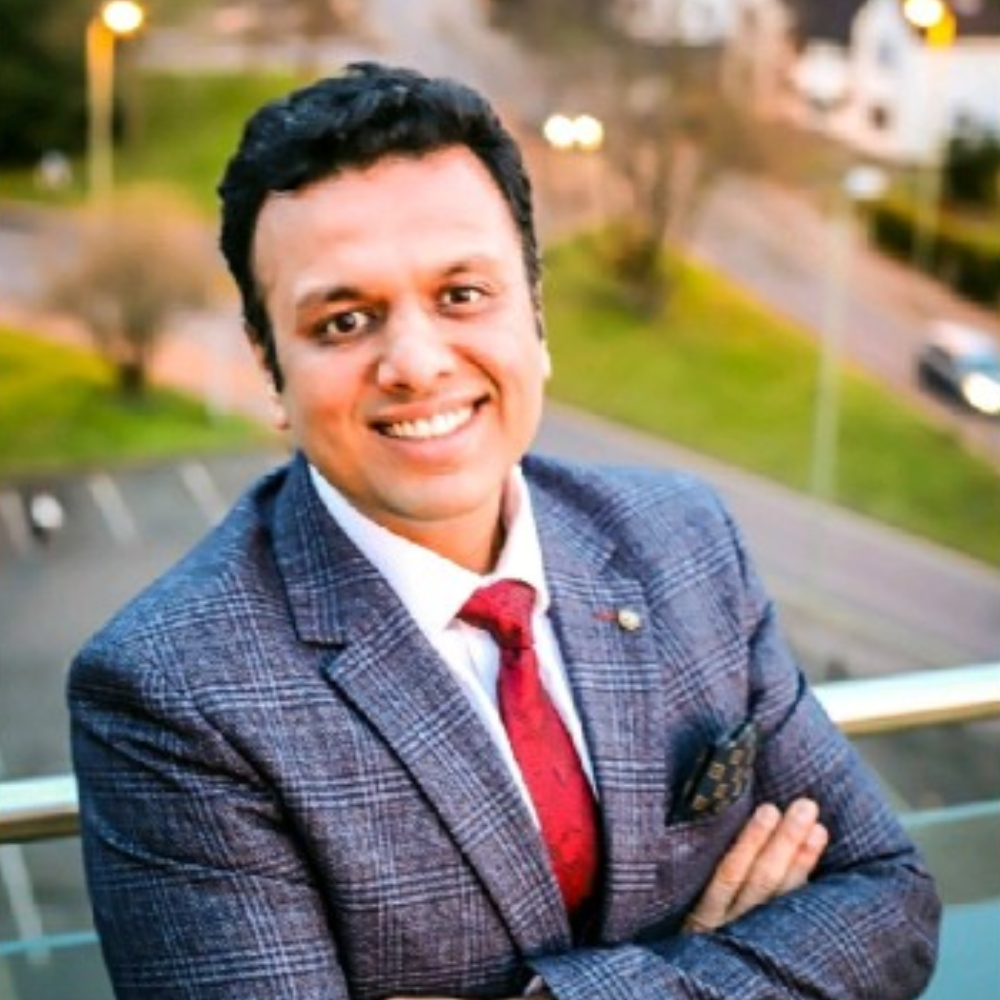 Smiling person in a gray suit and red tie, standing outdoors with arms crossed.