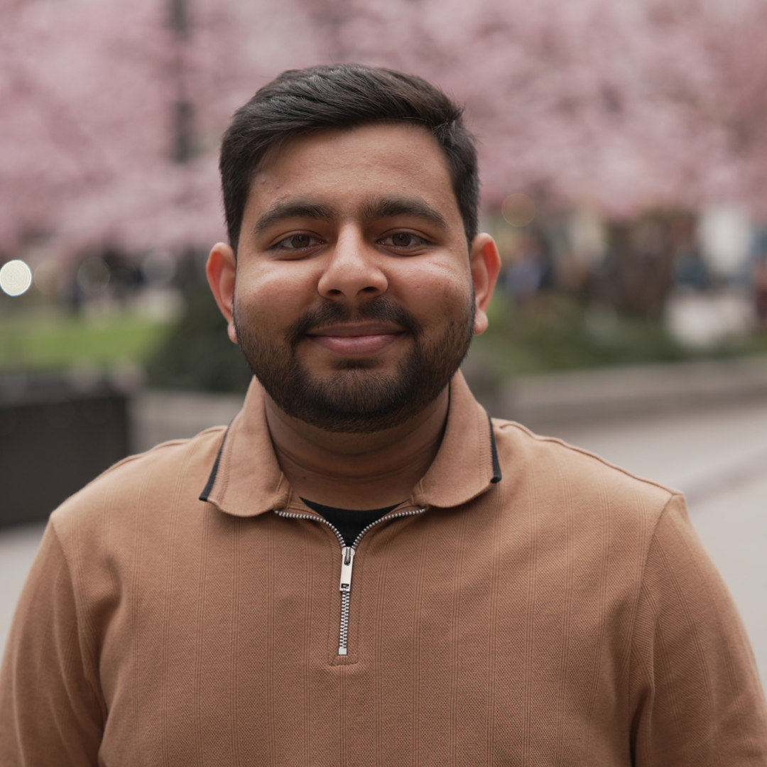 Smiling man in a tan sweater standing outdoors with pink cherry blossoms in the background