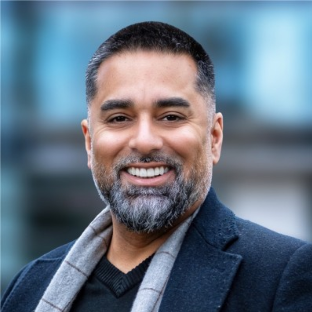 Smiling man with salt-and-pepper beard in dark blazer, standing outdoors with blurred blue background