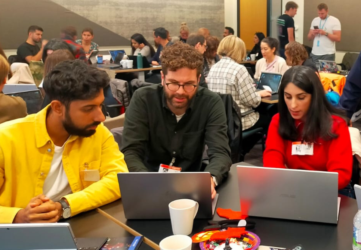 People working on laptops at a collaborative table in a busy indoor workspace, with coffee cups and notebooks.