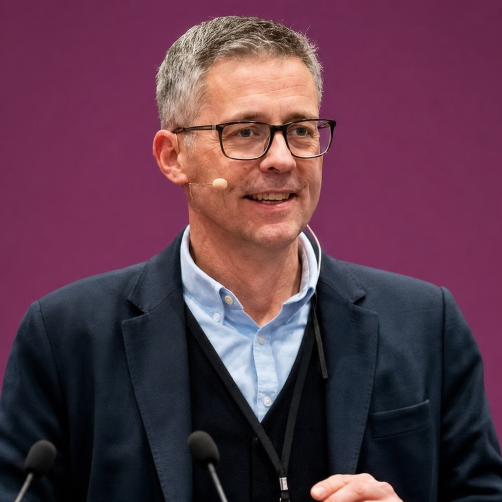 Man speaking at a podium with microphones, wearing glasses and a headset mic against a purple backdrop