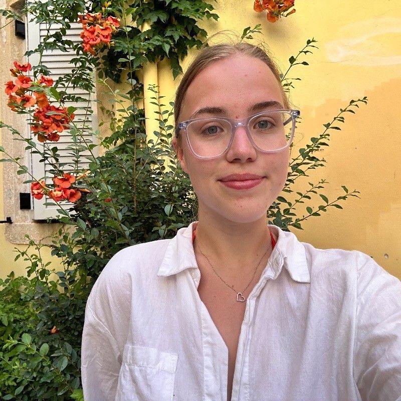 Person in a white shirt and glasses posing in front of a yellow wall and flowering vines.
