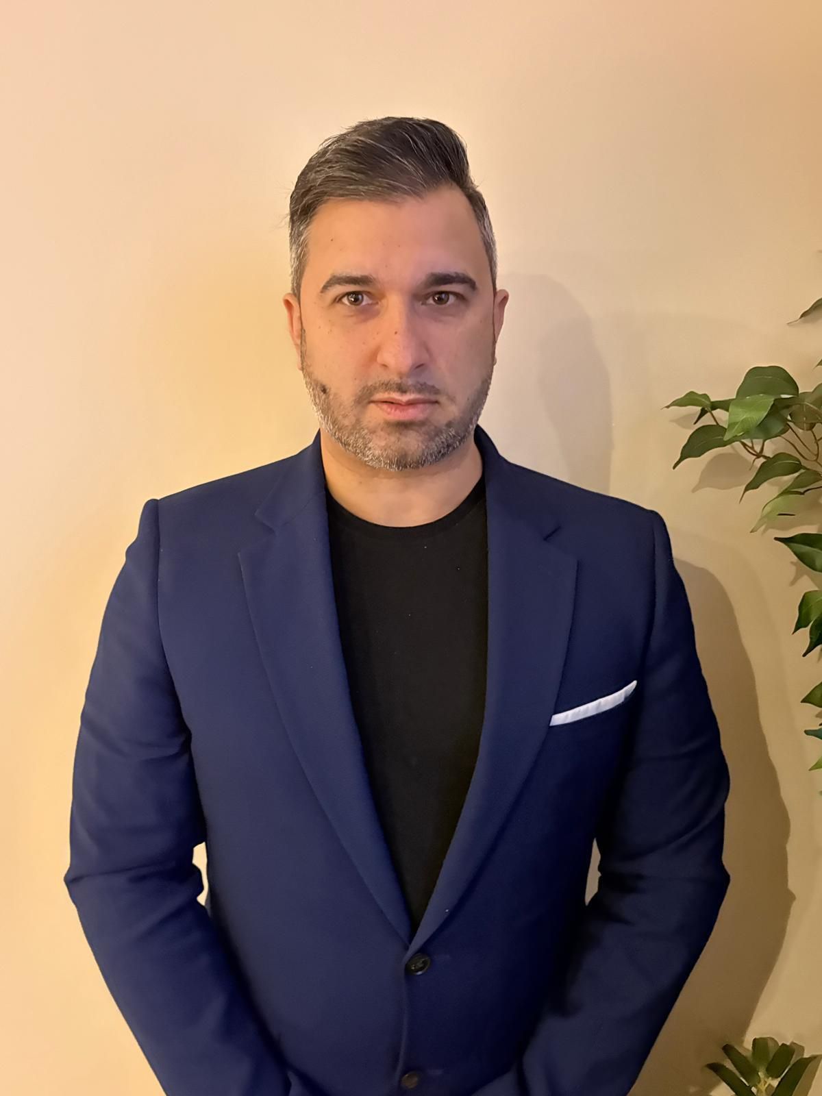 Man in a navy blazer and black shirt standing indoors against a beige wall with a potted plant nearby