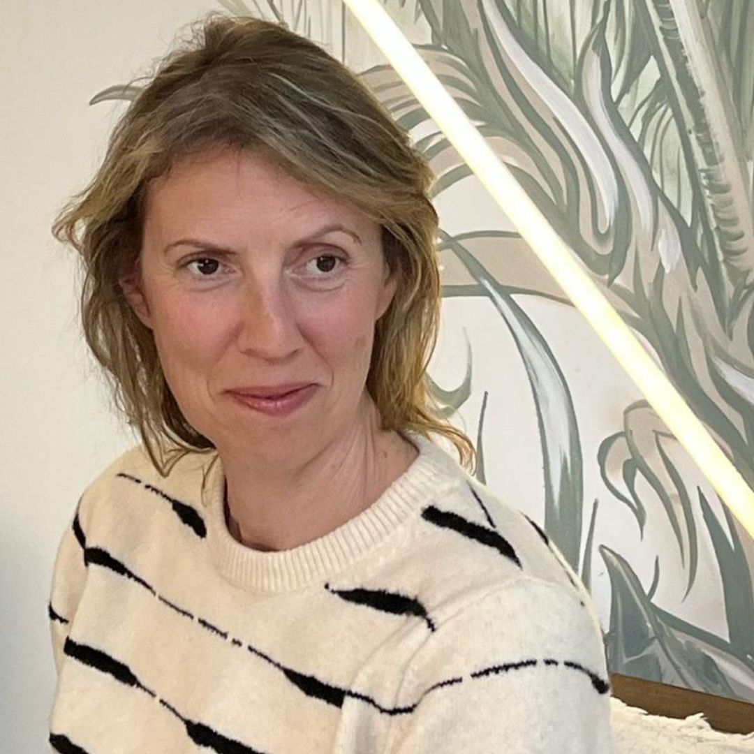 Woman in a cream sweater with black stripes, seated indoors against patterned wallpaper, slight smile.