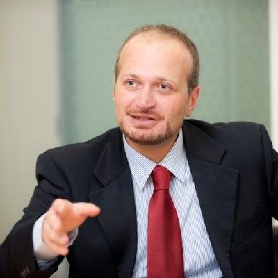 Man in a dark suit and red tie gestures while speaking in an office setting