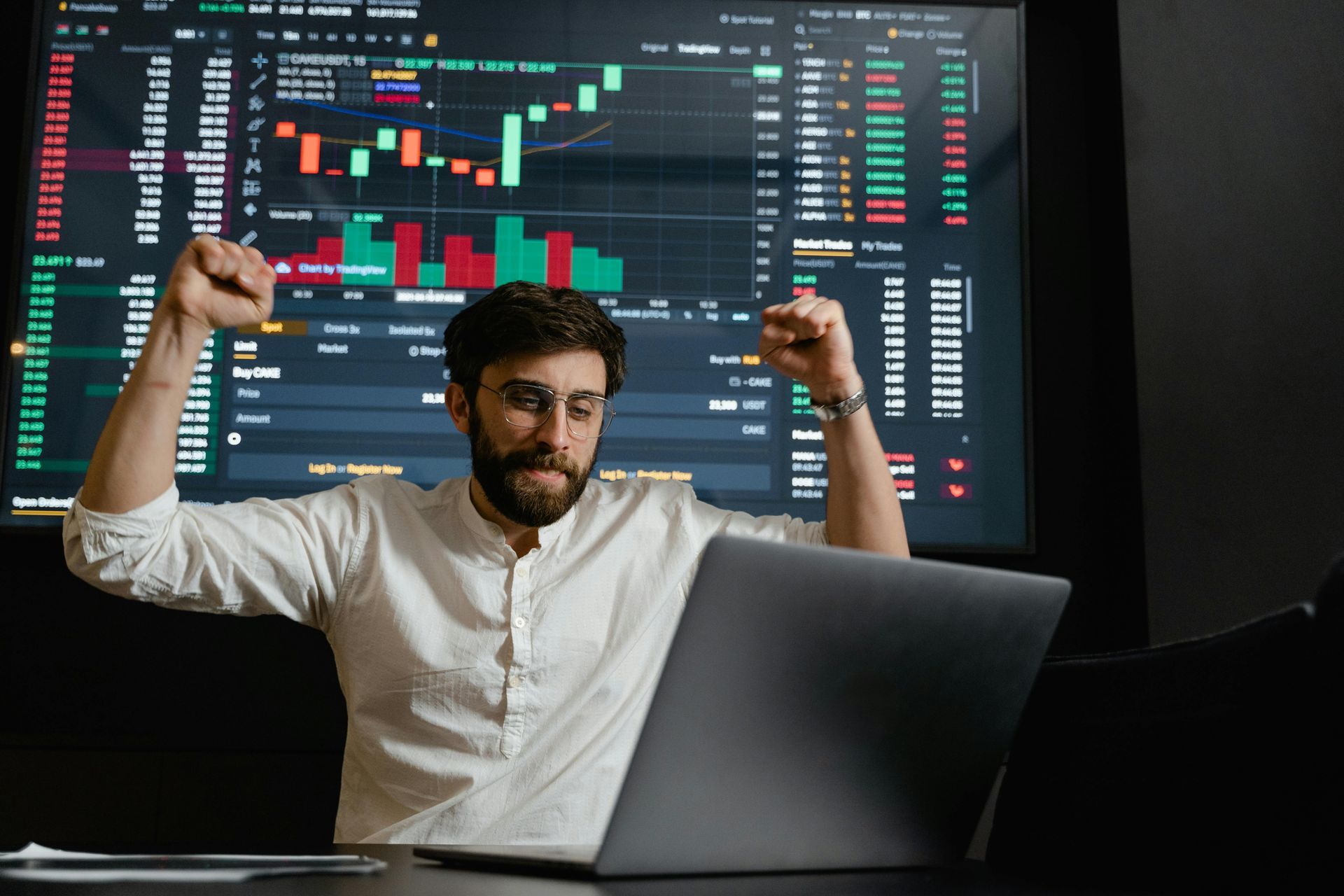 Trader celebrating with raised fists in front of a laptop and stock charts on multiple monitors