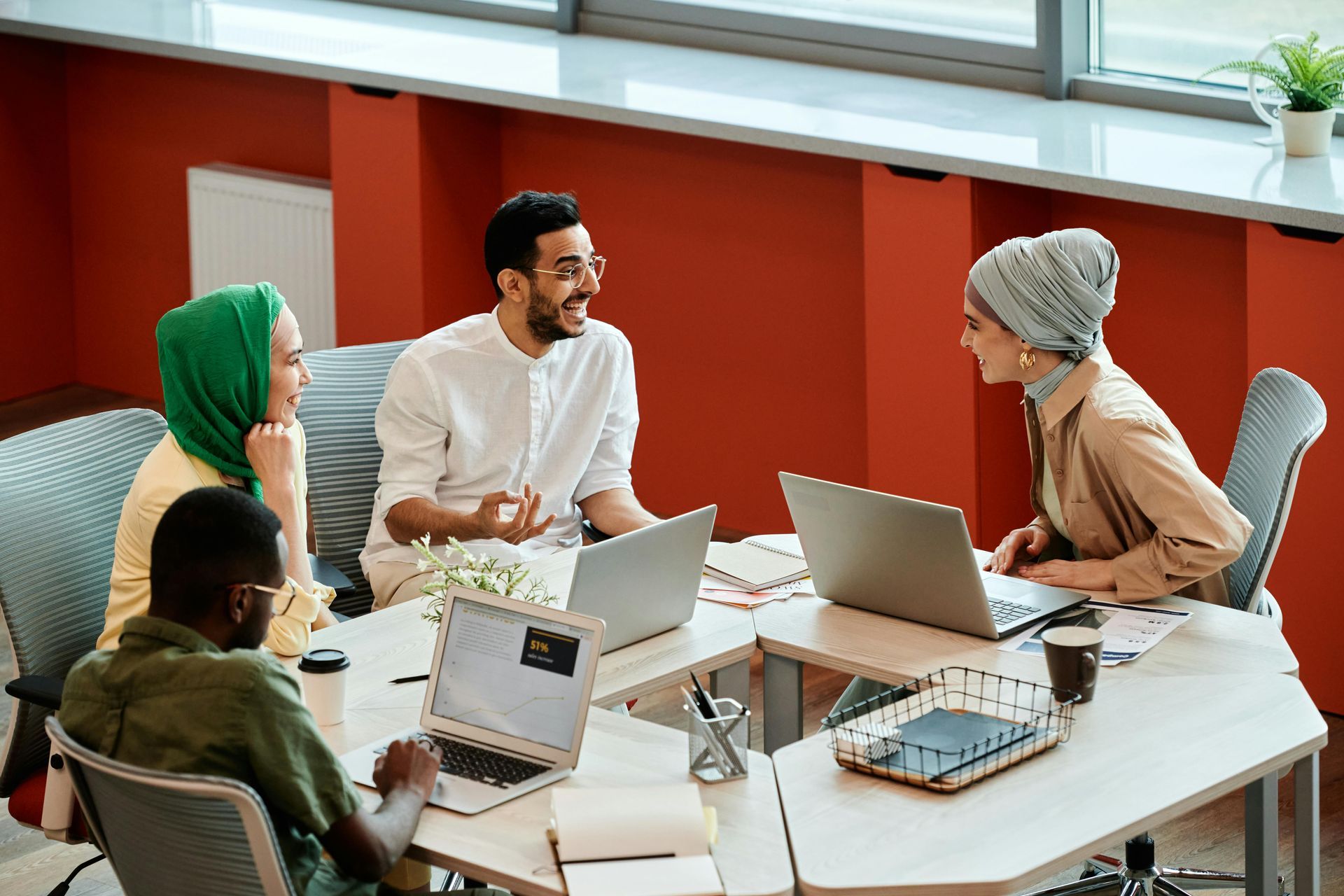 Four coworkers in a meeting around laptops at a bright conference table, talking in a modern office.