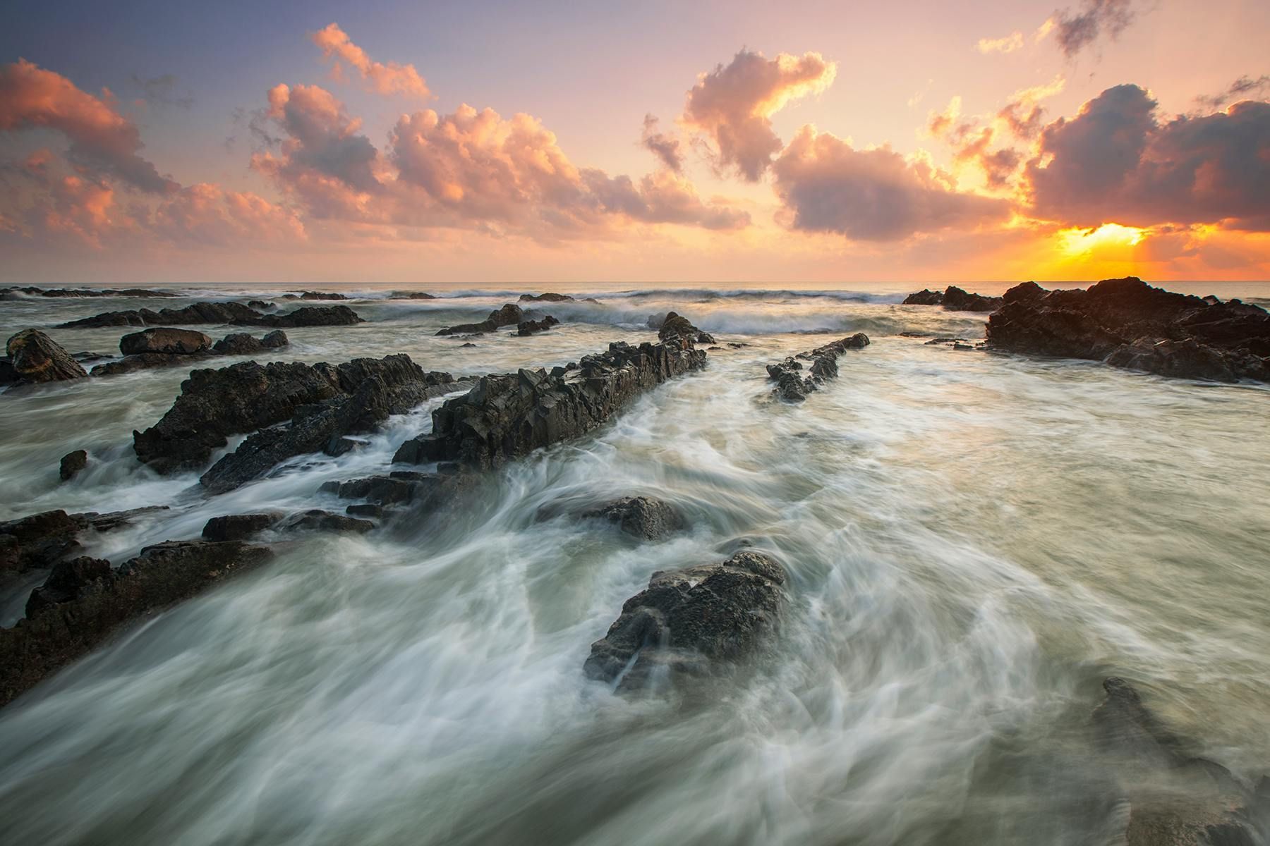 Sunset over rocky shoreline with waves rushing around dark rocks and pink clouds