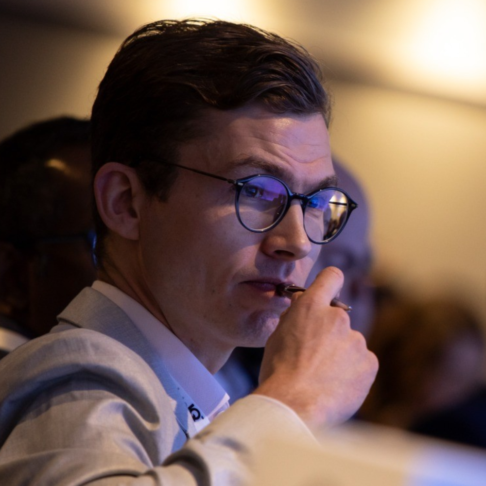 Man in glasses and light jacket resting his hand on his face in a dimly lit audience setting