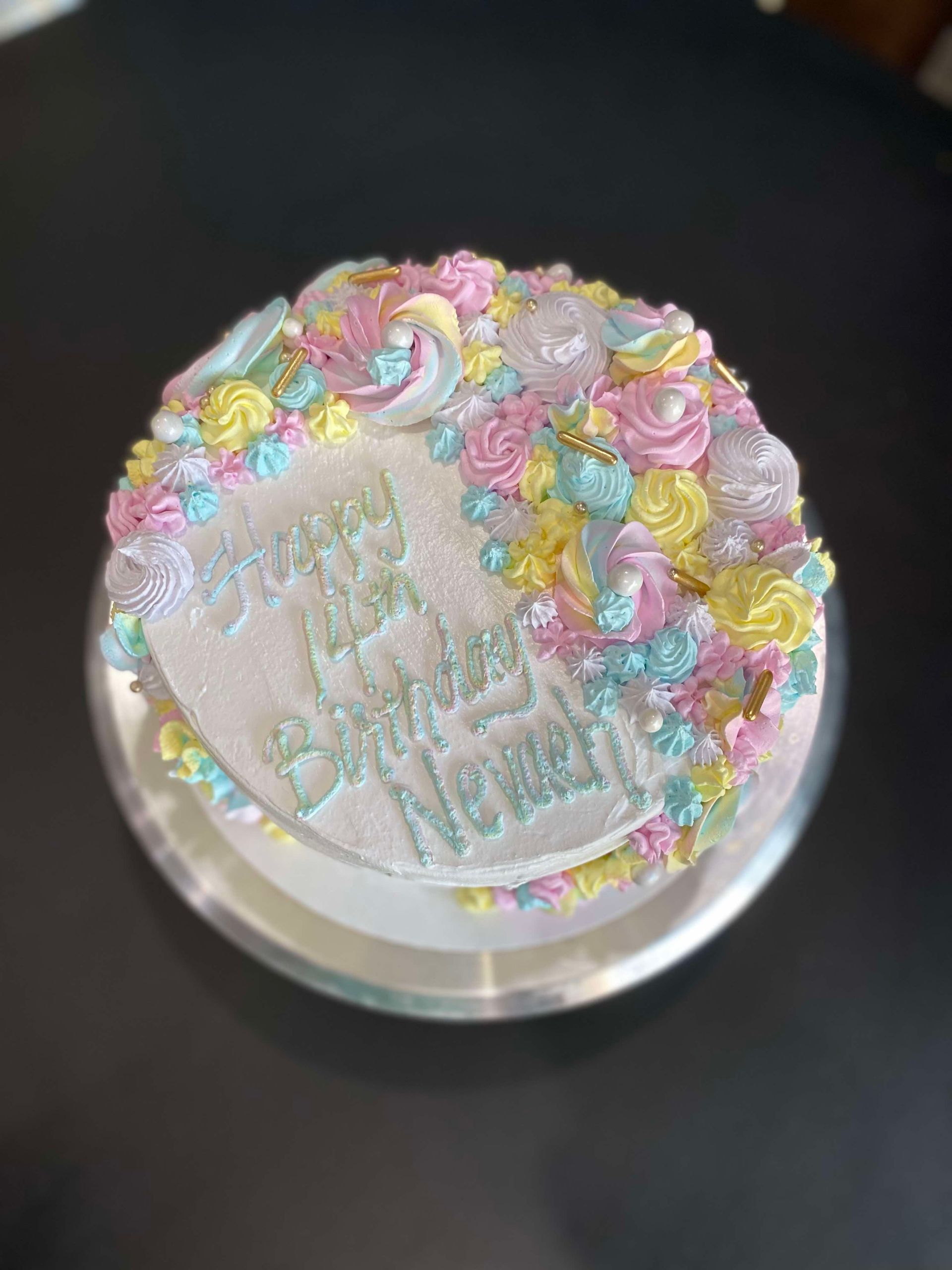 A round birthday cake with white frosting, decorated with pastel pink, blue, and yellow piped rosettes and gold sprinkles.