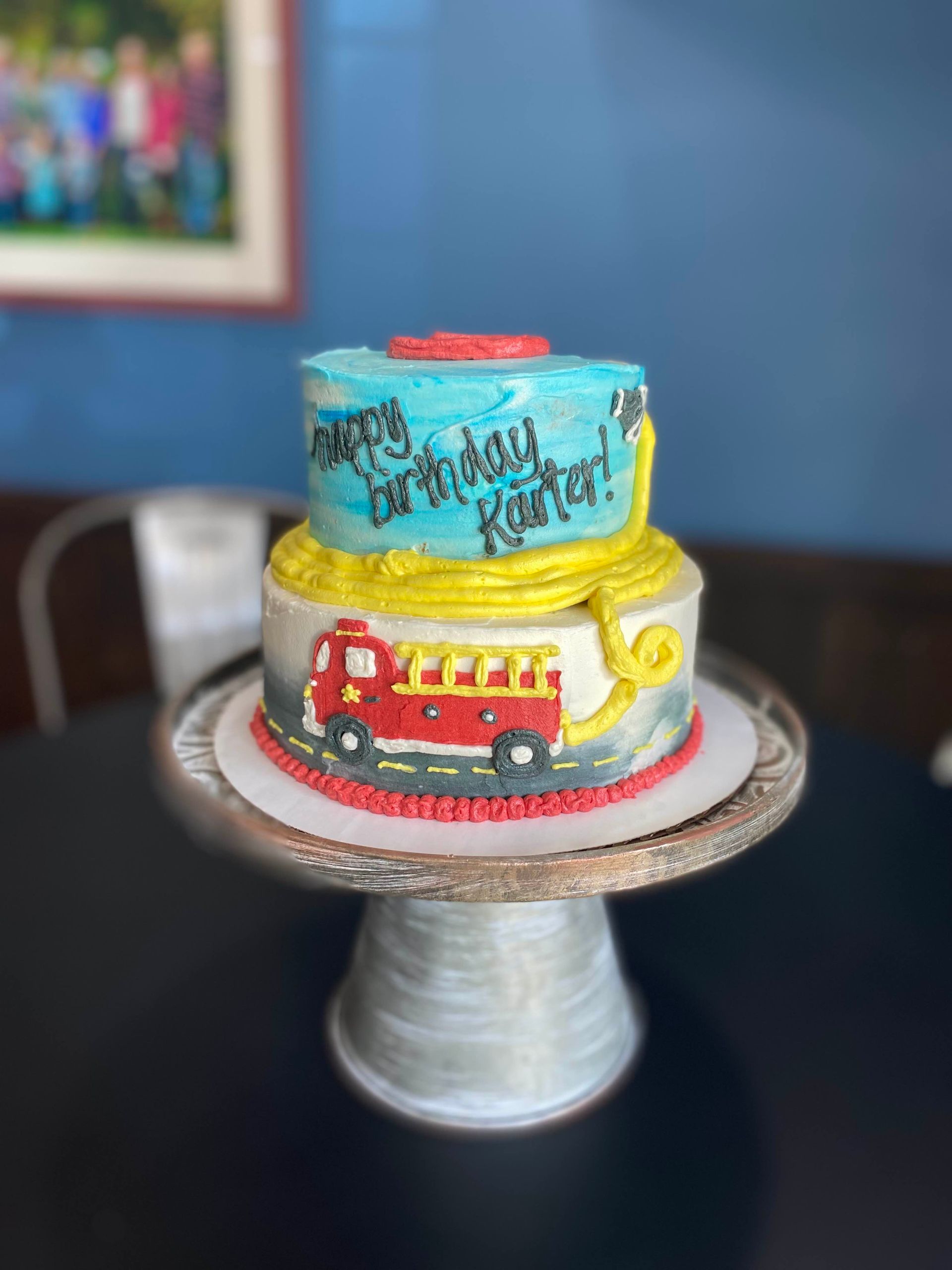 A two-tier birthday cake decorated with a fire truck and a yellow hose on a silver cake stand against a blue wall.