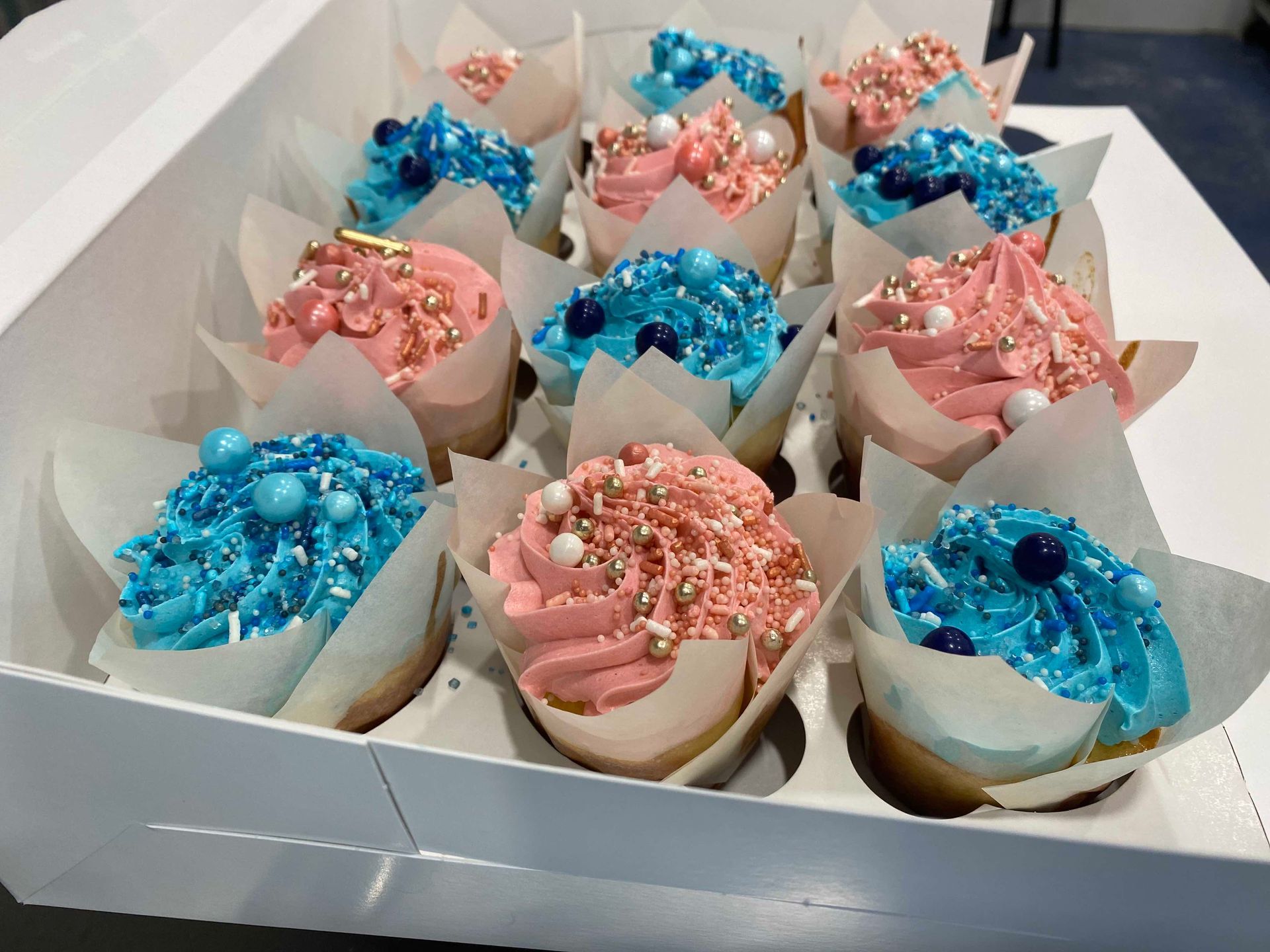 A dozen cupcakes in a white box, featuring alternating pink and blue frosting decorated with sprinkles and sugar pearls.