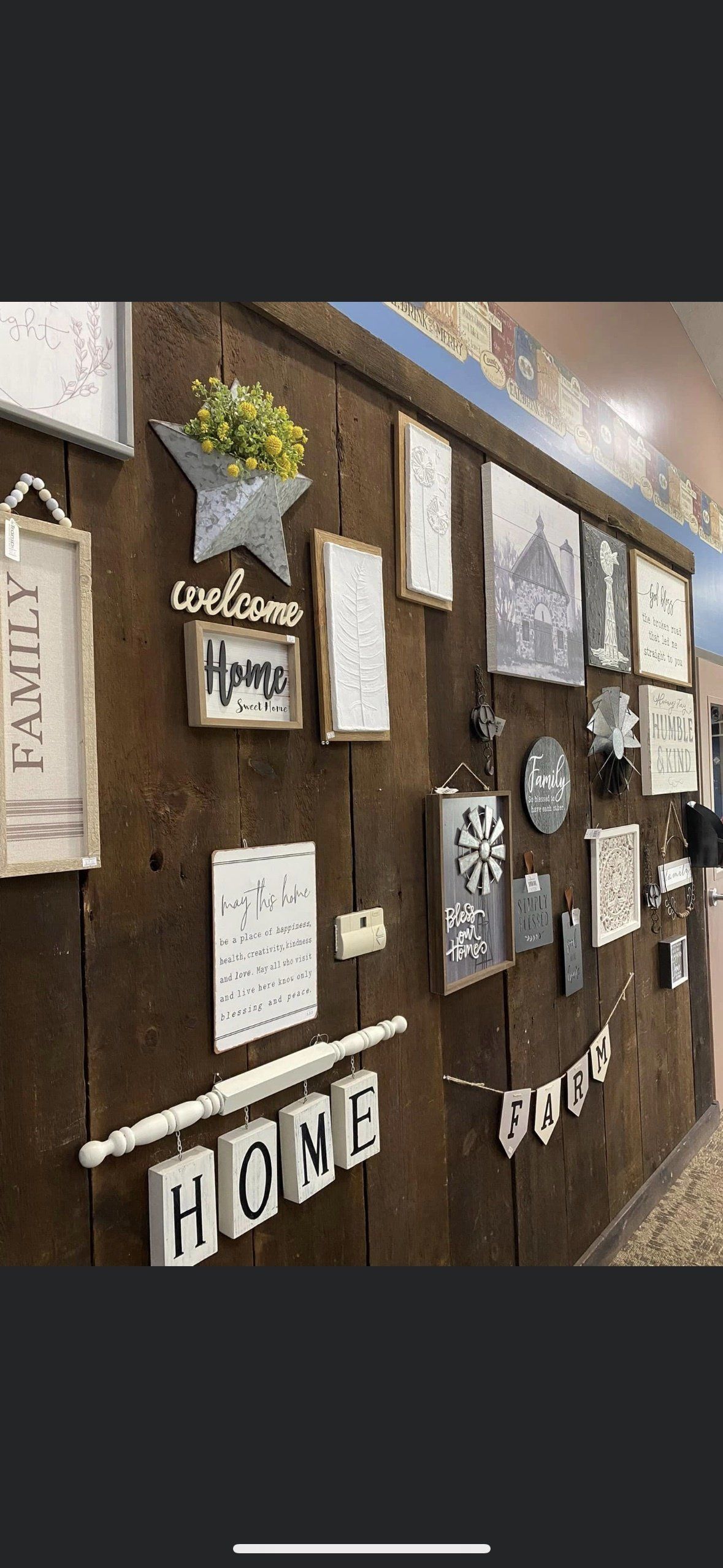 A rustic, dark-wood wall display featuring various farmhouse-style signs, decorative frames, and home-themed accents.