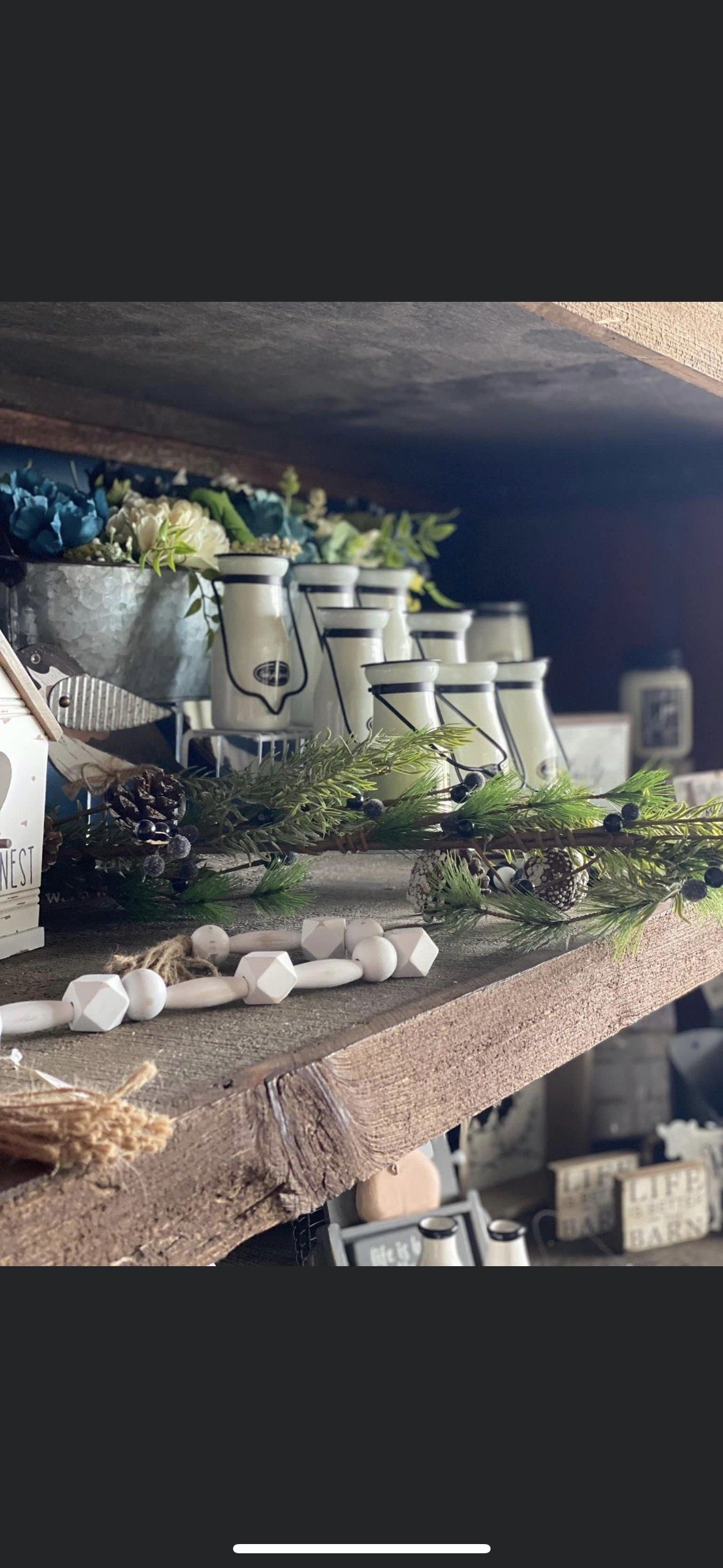 A rustic wooden shelf is decorated with a collection of white pitchers, greenery, and a string of white wooden beads.