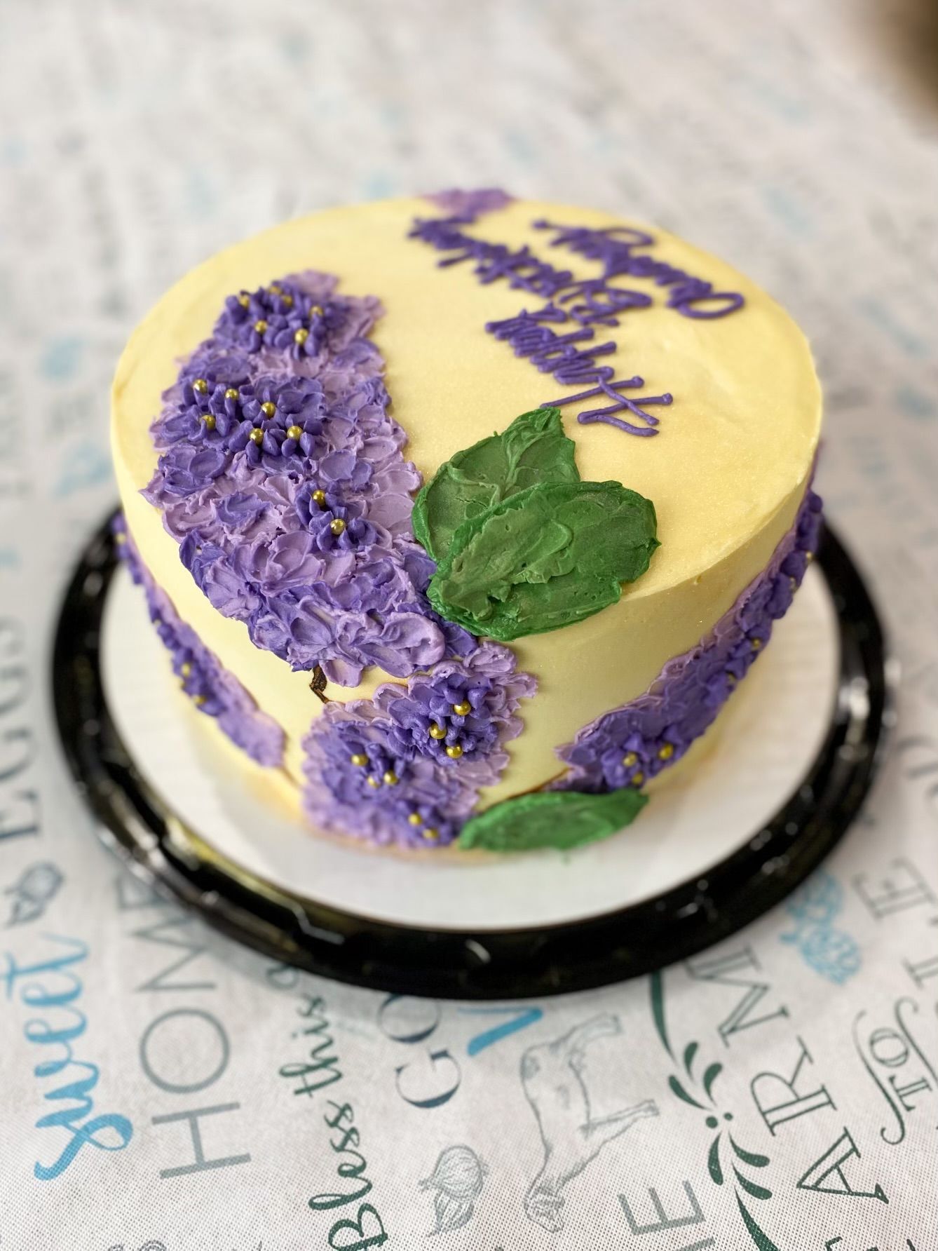A yellow frosted cake decorated with purple piped flower clusters and green frosting leaves, reading