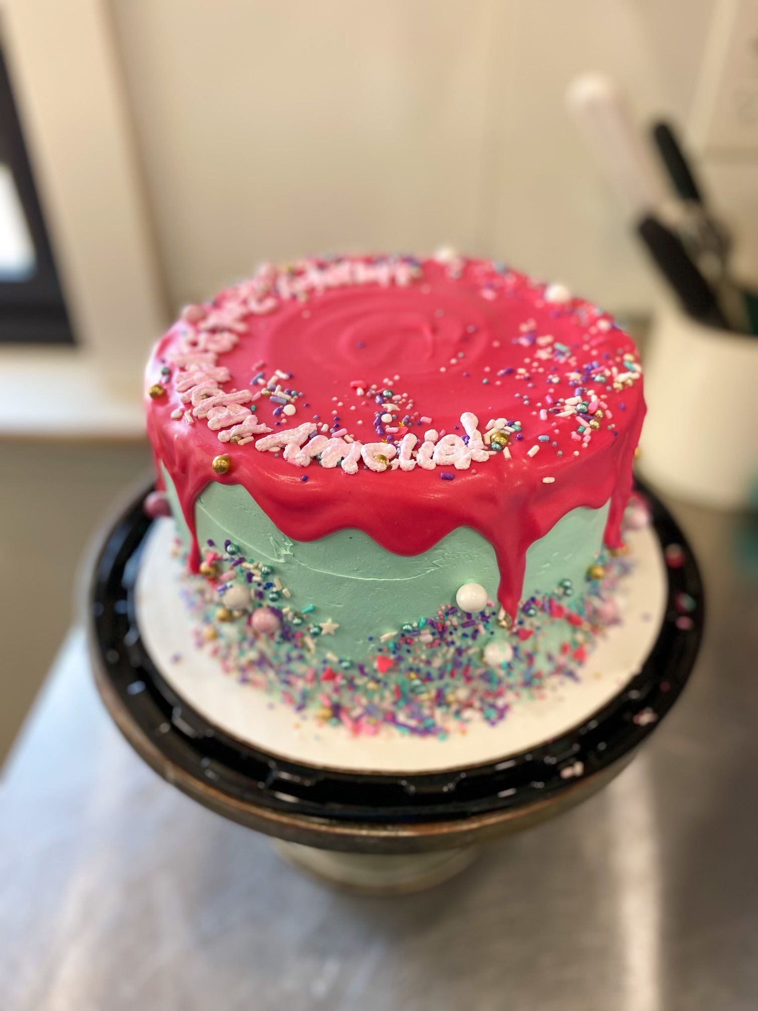 A light blue cake with a pink drip glaze, topped with sprinkles and white piped lettering on a dark cake stand.