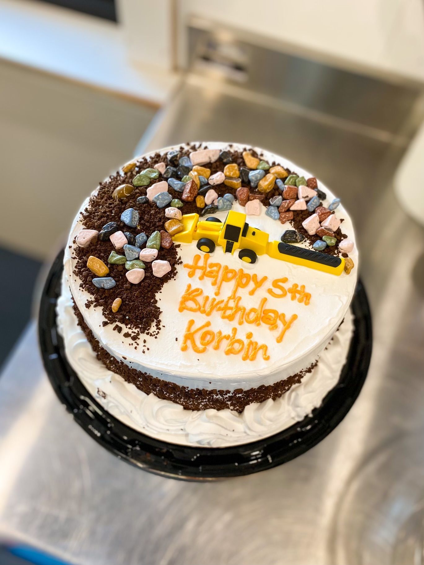 A round birthday cake with white frosting, a yellow toy construction truck, chocolate sprinkles, and candy rock toppings.