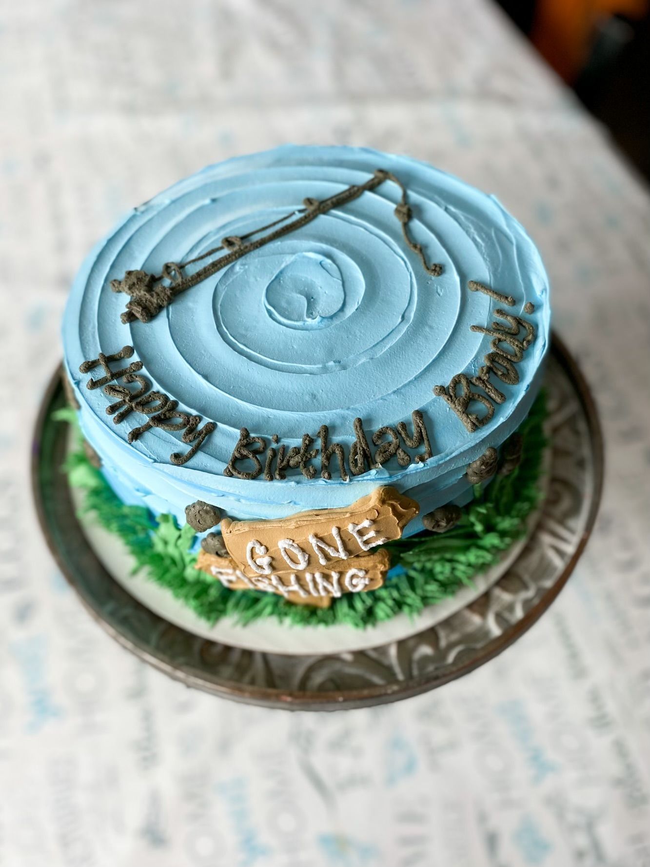 A round, blue-frosted birthday cake topped with a fishing pole, saying