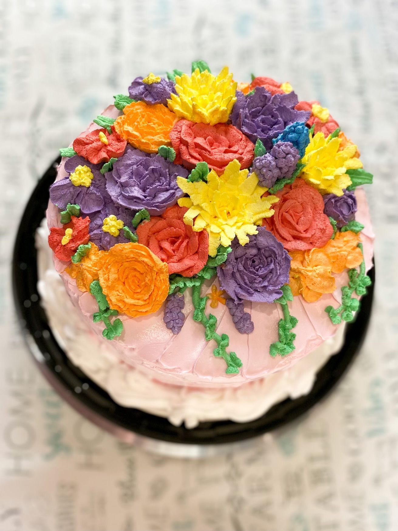 A pink frosted cake topped with a vibrant arrangement of yellow, orange, red, and purple buttercream flowers.