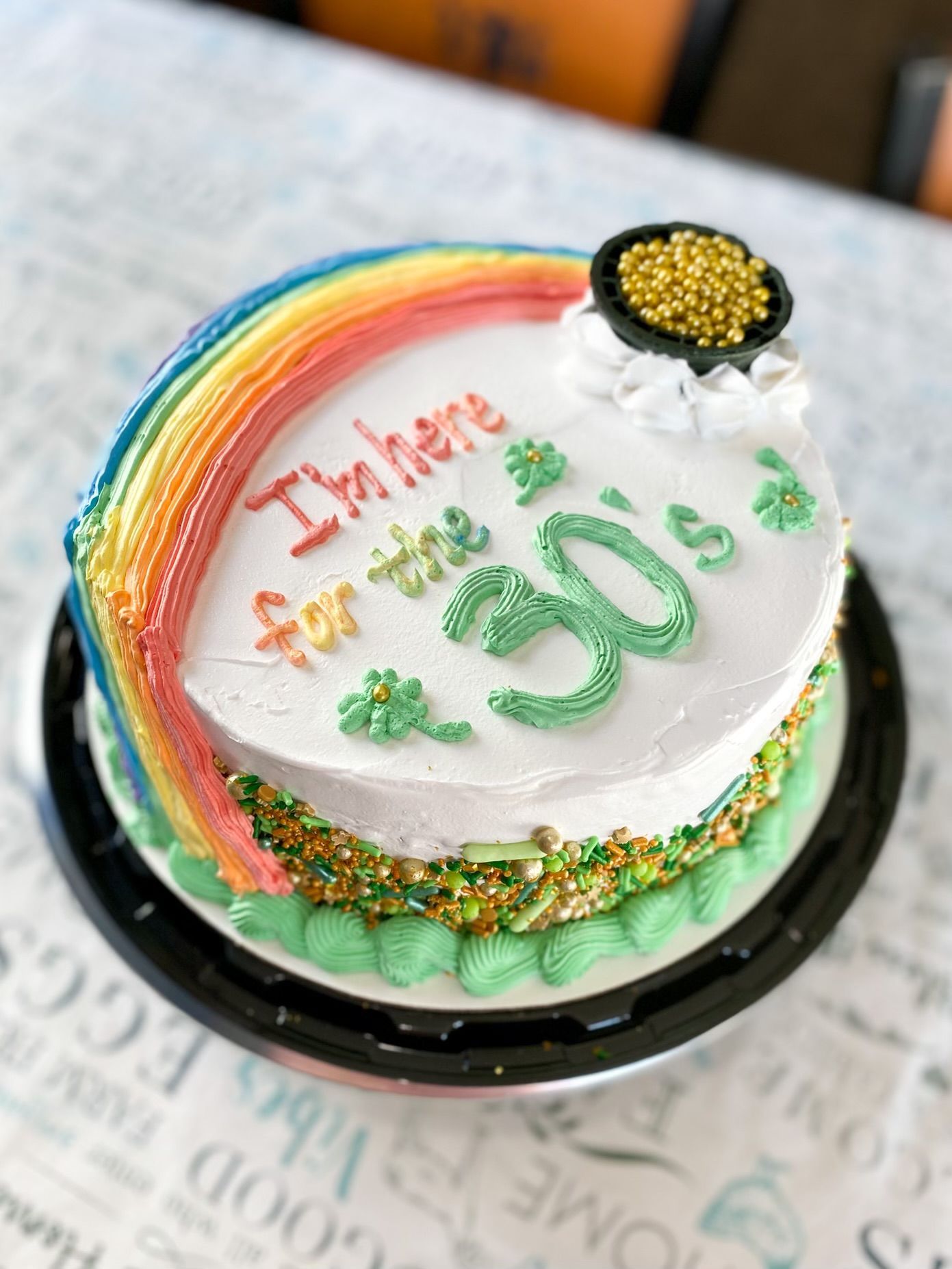 A round birthday cake with a rainbow, shamrocks, and a pot of gold, inscribed with
