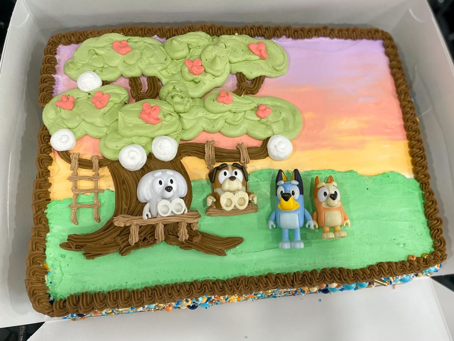A rectangular cake featuring a buttercream tree, a ladder, and Bluey and Bingo figures standing on a green lawn background.