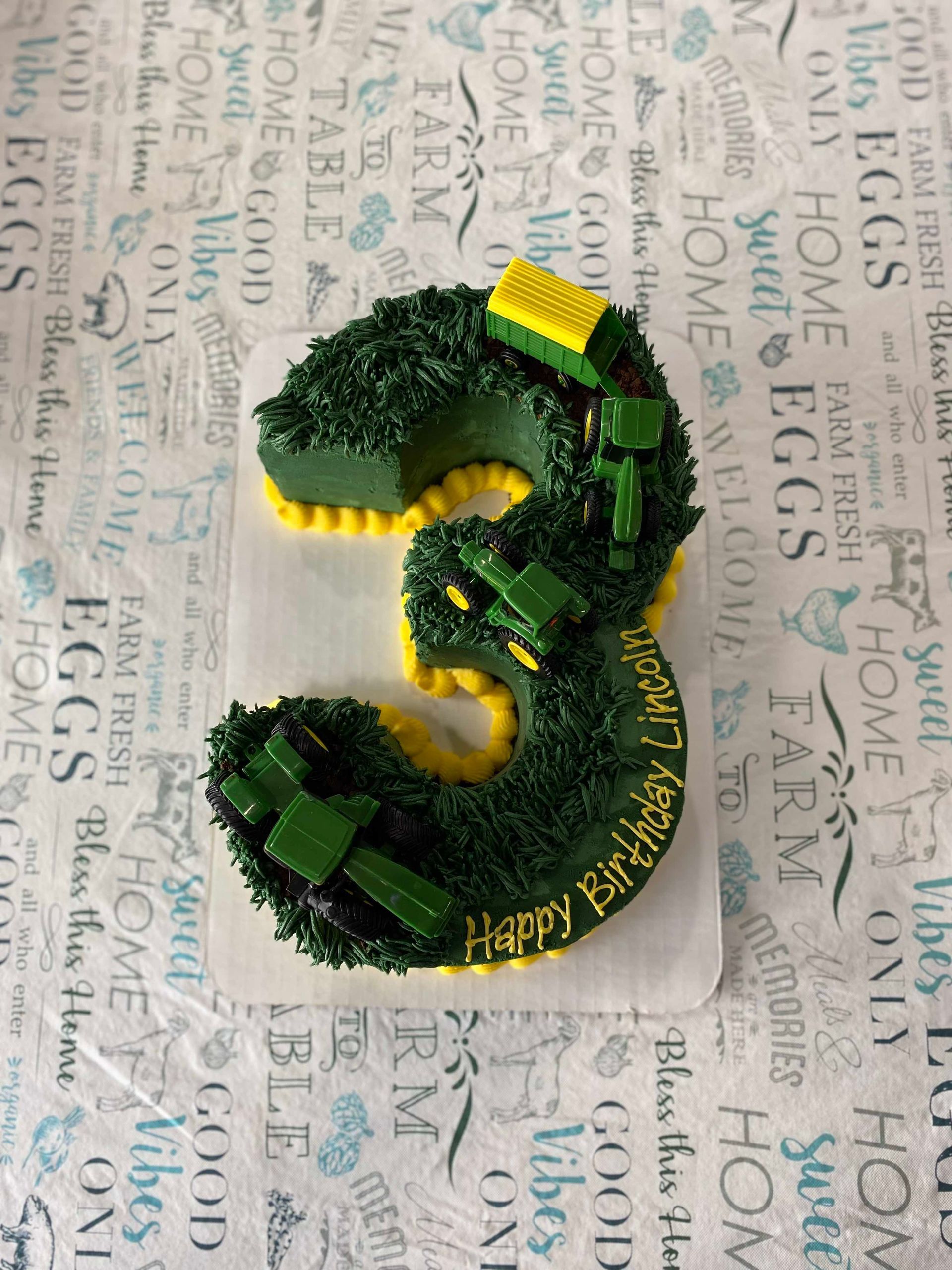 A number 3-shaped birthday cake, frosted in dark green grass texture, decorated with toy John Deere tractors on top.