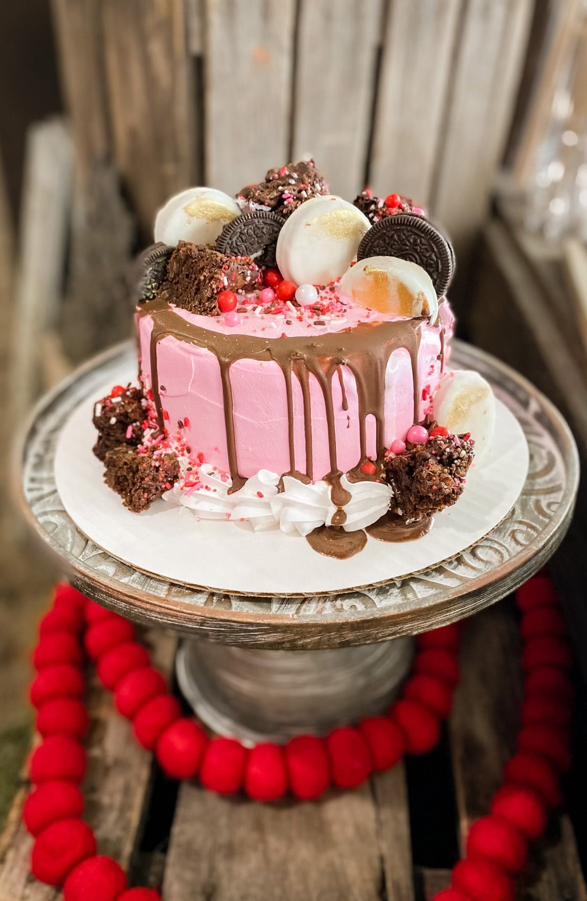 A pink frosted cake with a chocolate drip, topped with cookies and brownie bites, on a stand with a red garland base.
