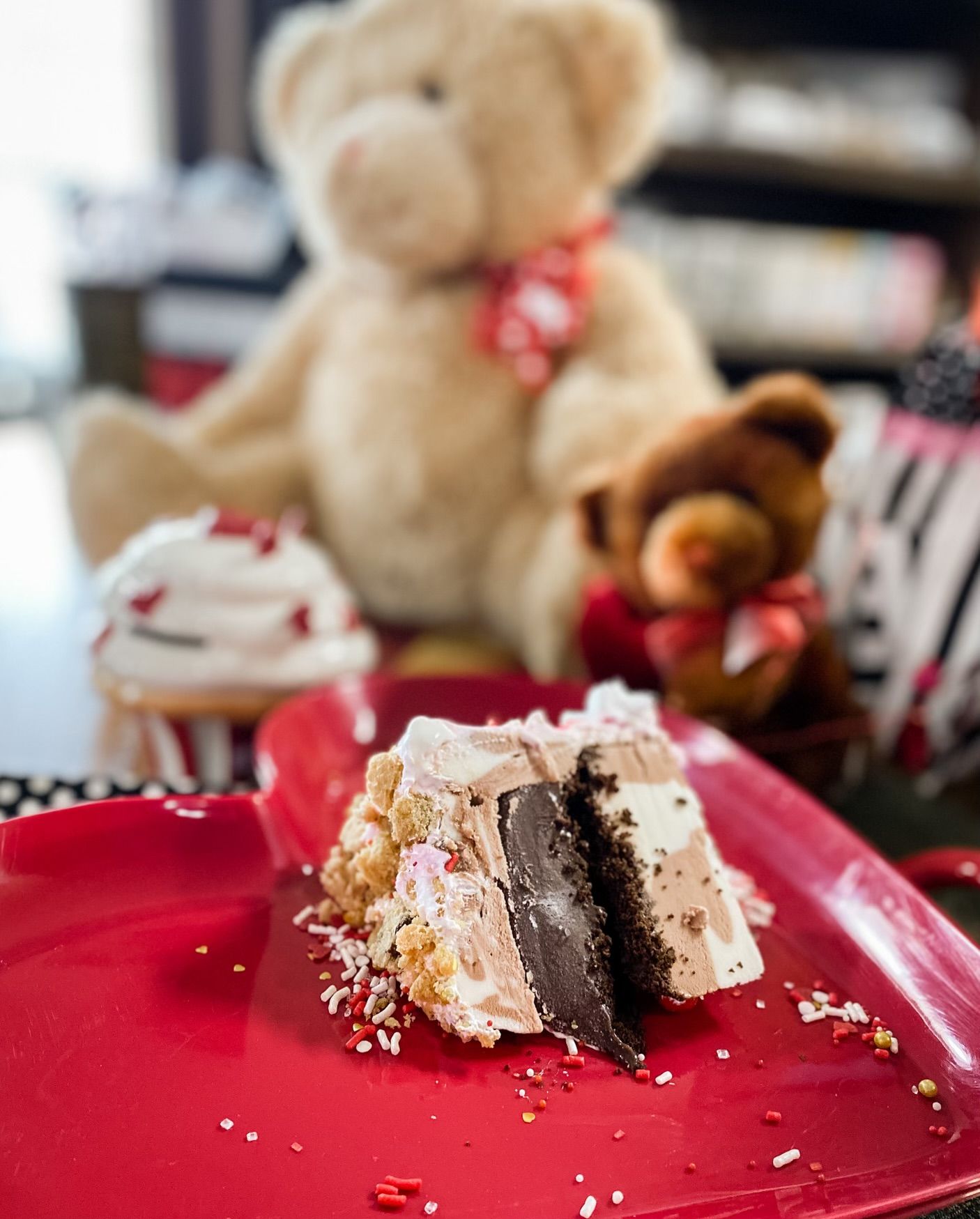 A slice of chocolate cake topped with frosting and sprinkles on a red heart-shaped plate, with teddy bears in the back.