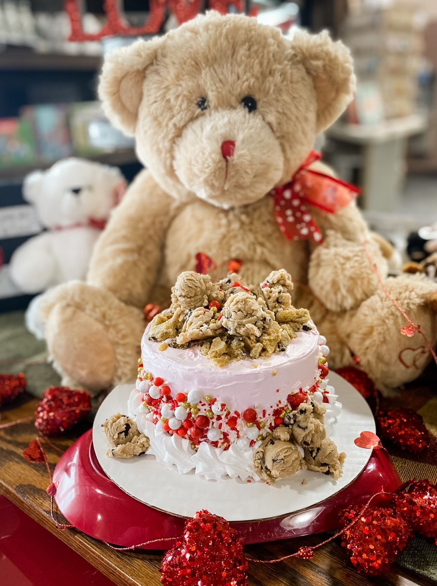 A small, pink-frosted cake topped with cookie dough, sitting on a table with teddy bears and red heart decorations.