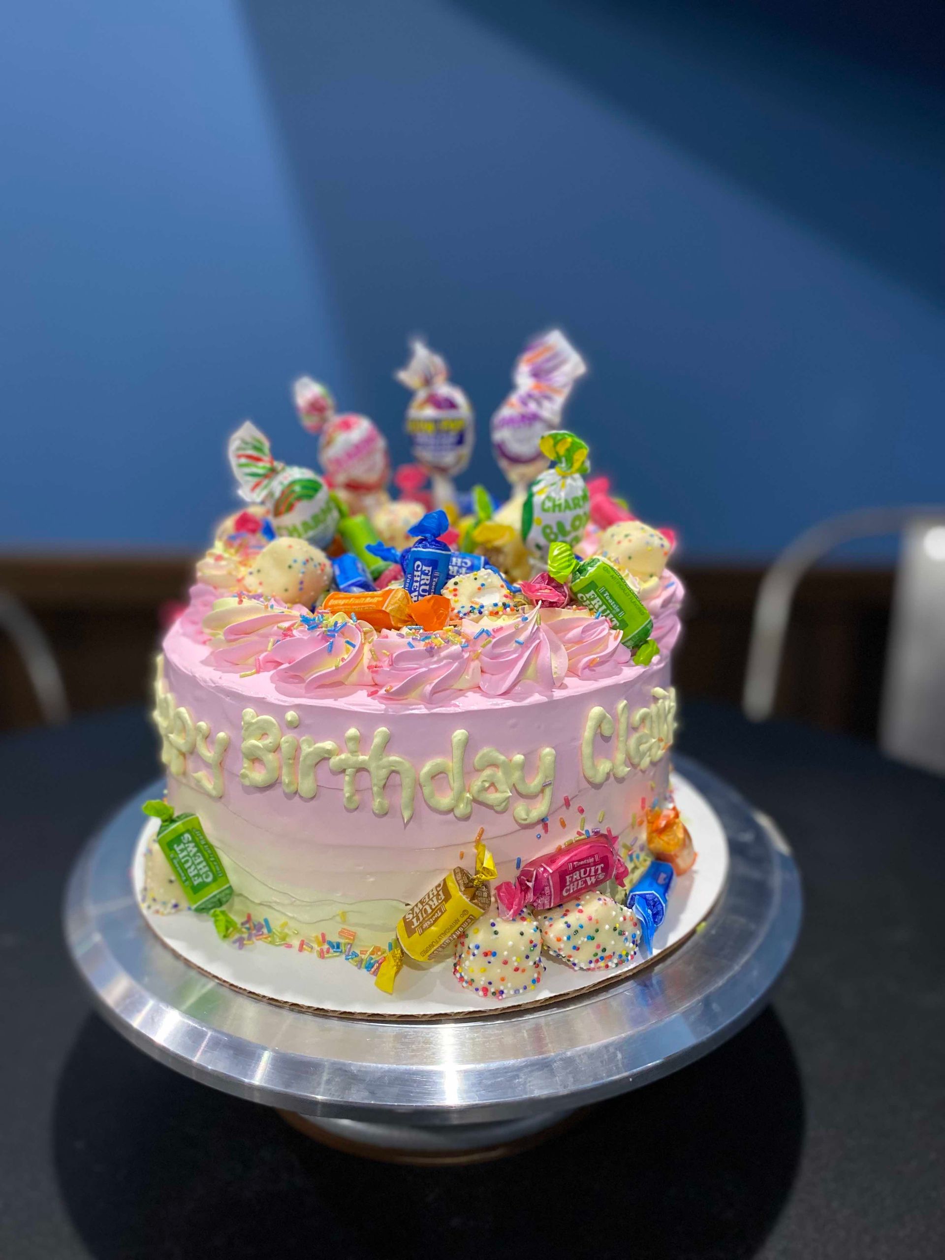 A round birthday cake with pink-to-white ombre frosting, topped with various colorful candies on a silver cake stand.