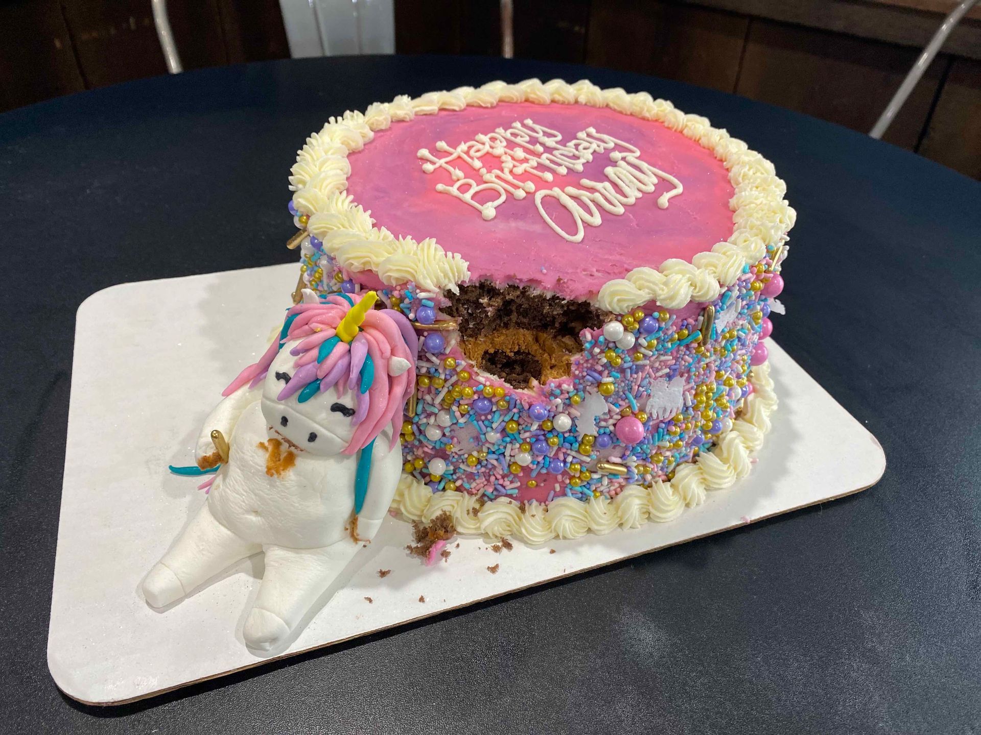 A birthday cake decorated with pink icing, sprinkles, and a white unicorn figurine appearing to eat a slice from the side.