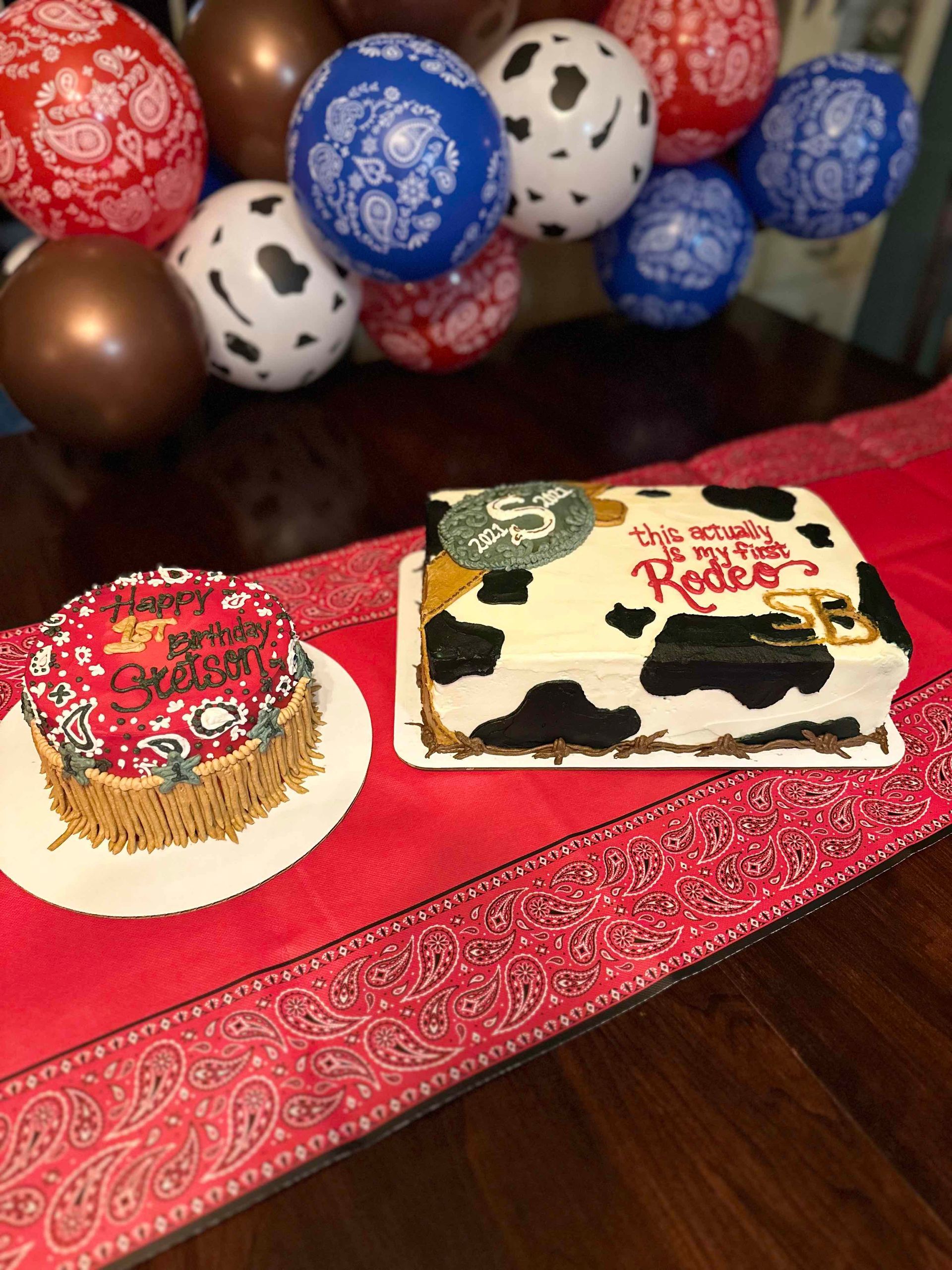 Two cowboy-themed cakes with cow print and red bandana patterns sit on a table with matching balloons in the background.