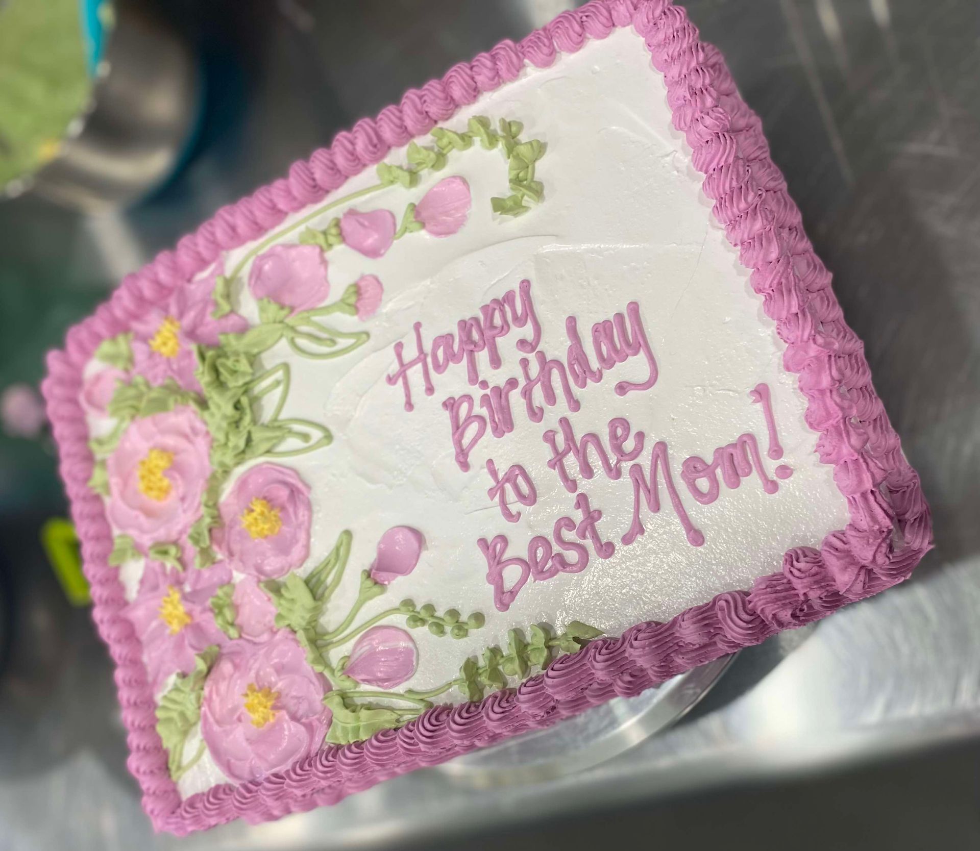A rectangular white cake with purple piped borders, decorated with green vines and pink flowers, inscribed
