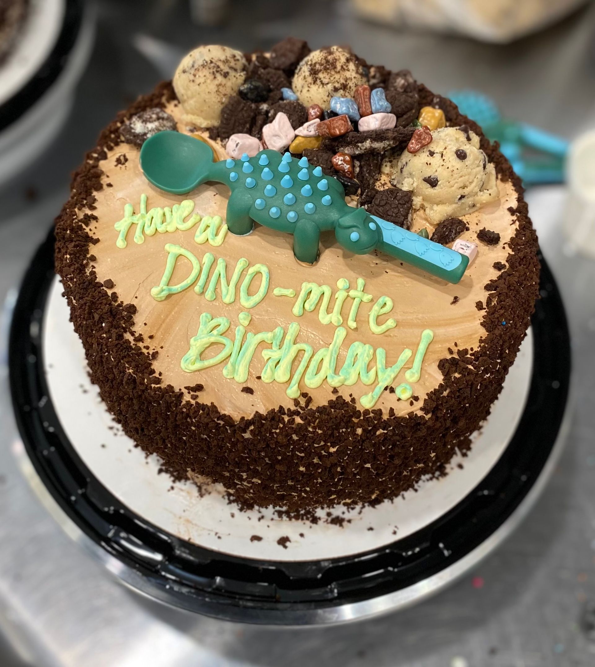 A round birthday cake with brown frosting and cookie crumbs, topped with a green toy dinosaur, spoon, and cookie dough.