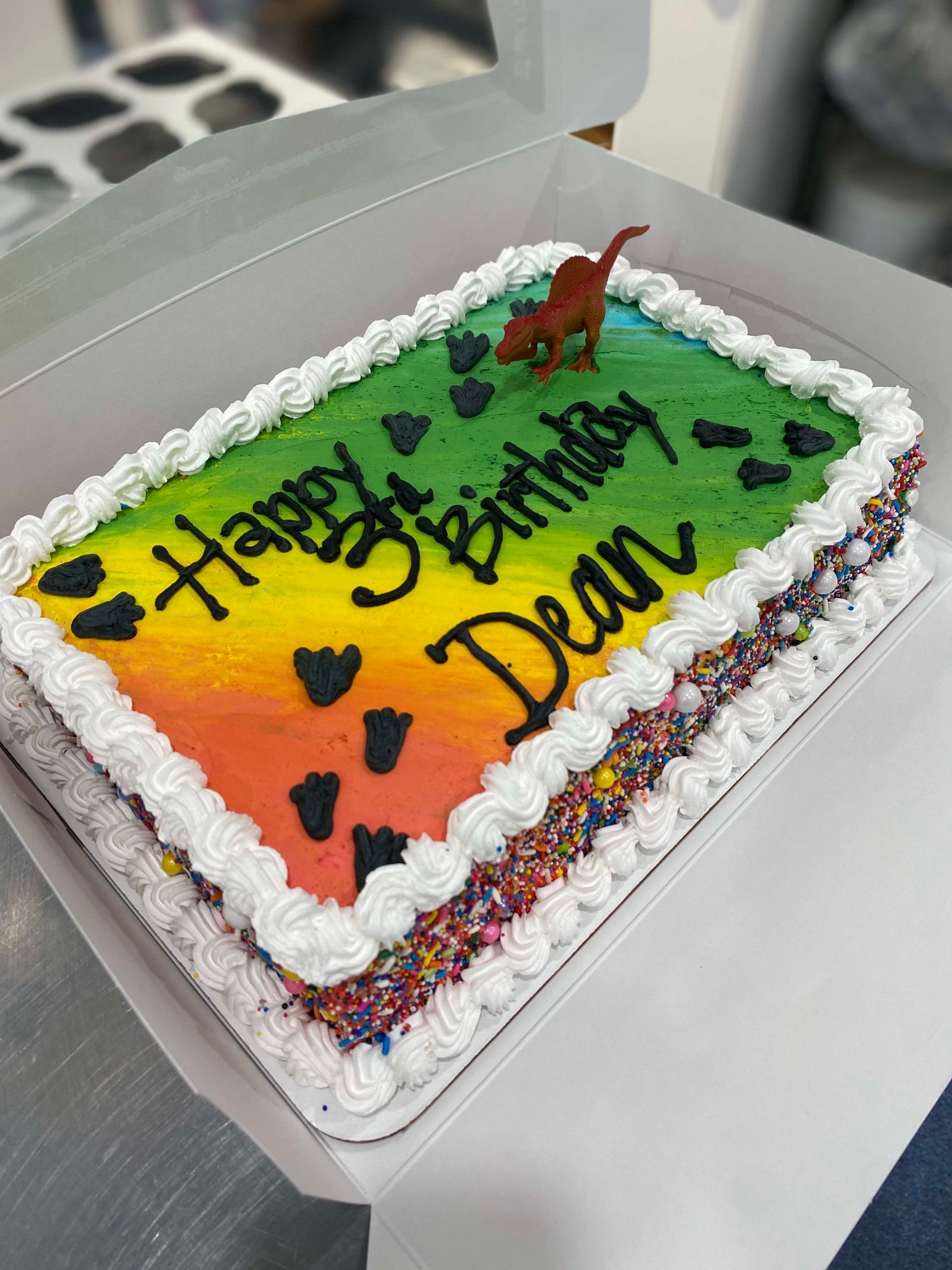Rectangular sheet cake with rainbow-colored frosting, dinosaur footprints, a toy dinosaur, and