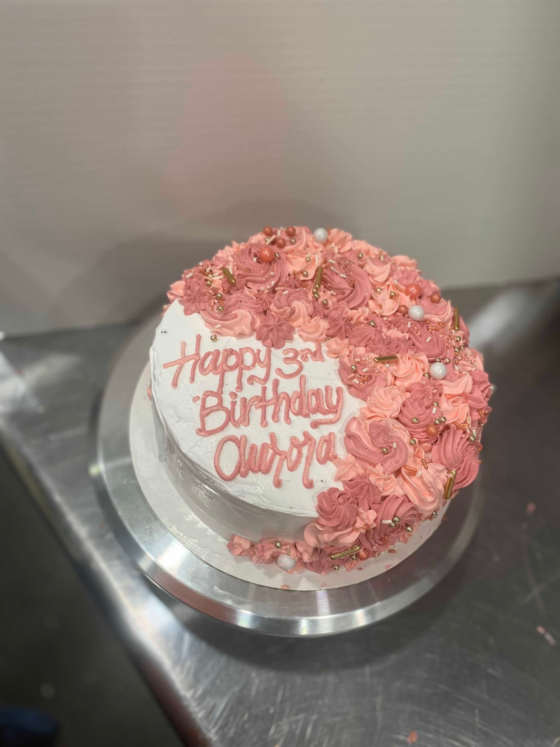 A white-frosted birthday cake topped with pink floral piping, gold accents, and white pearls, labeled
