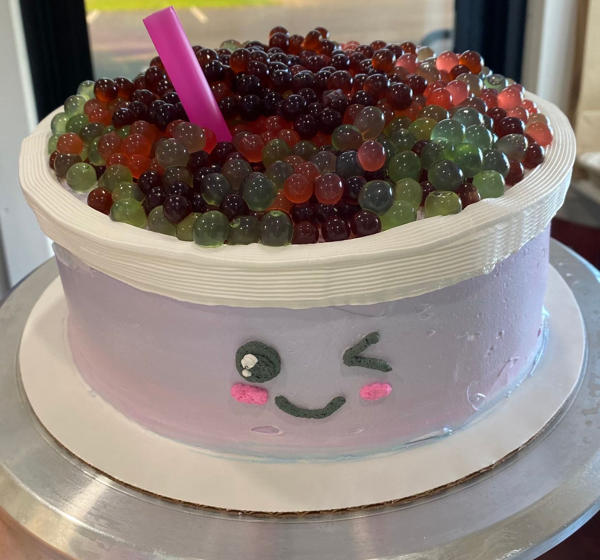 A round cake decorated to look like bubble tea, with a light purple face, pink blush, and colorful boba pearls on top.