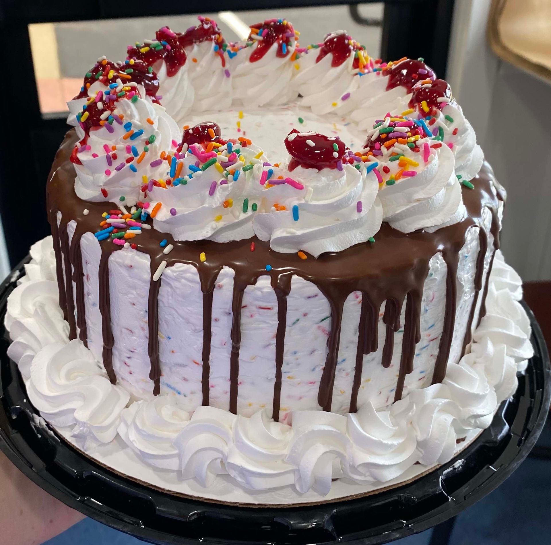 A round confetti cake decorated with chocolate drip, whipped cream rosettes, sprinkles, and dollops of red jam.
