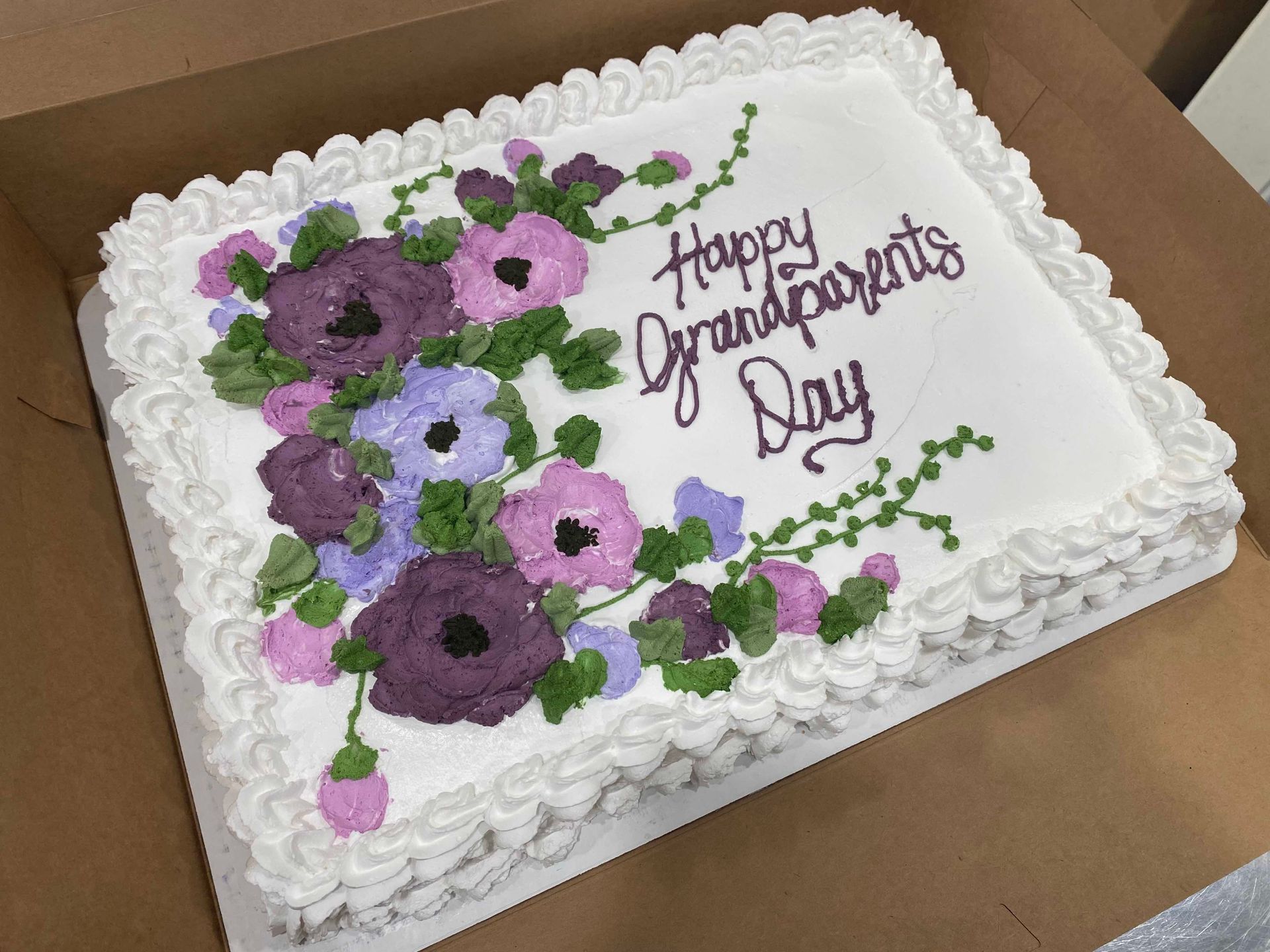 A rectangular cake in a cardboard box decorated with purple and lavender frosting flowers and