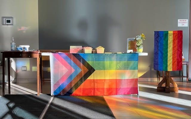 Photo of our sunlit foyer, with two pride flags hanging on the welcome table and lectern.