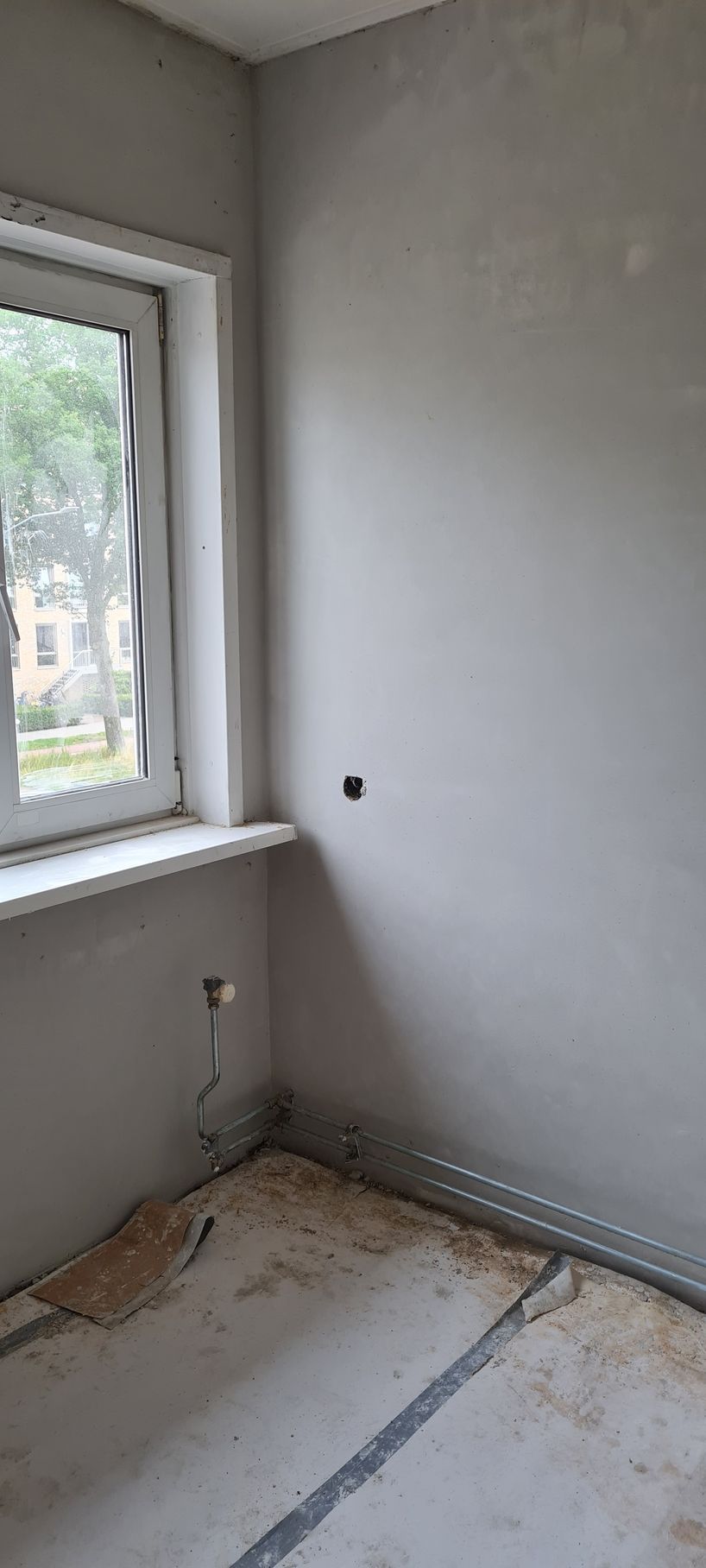 Een kleine, onafgewerkte kamer met een raam, kale muren, blootliggende draden en puin op de vloer.