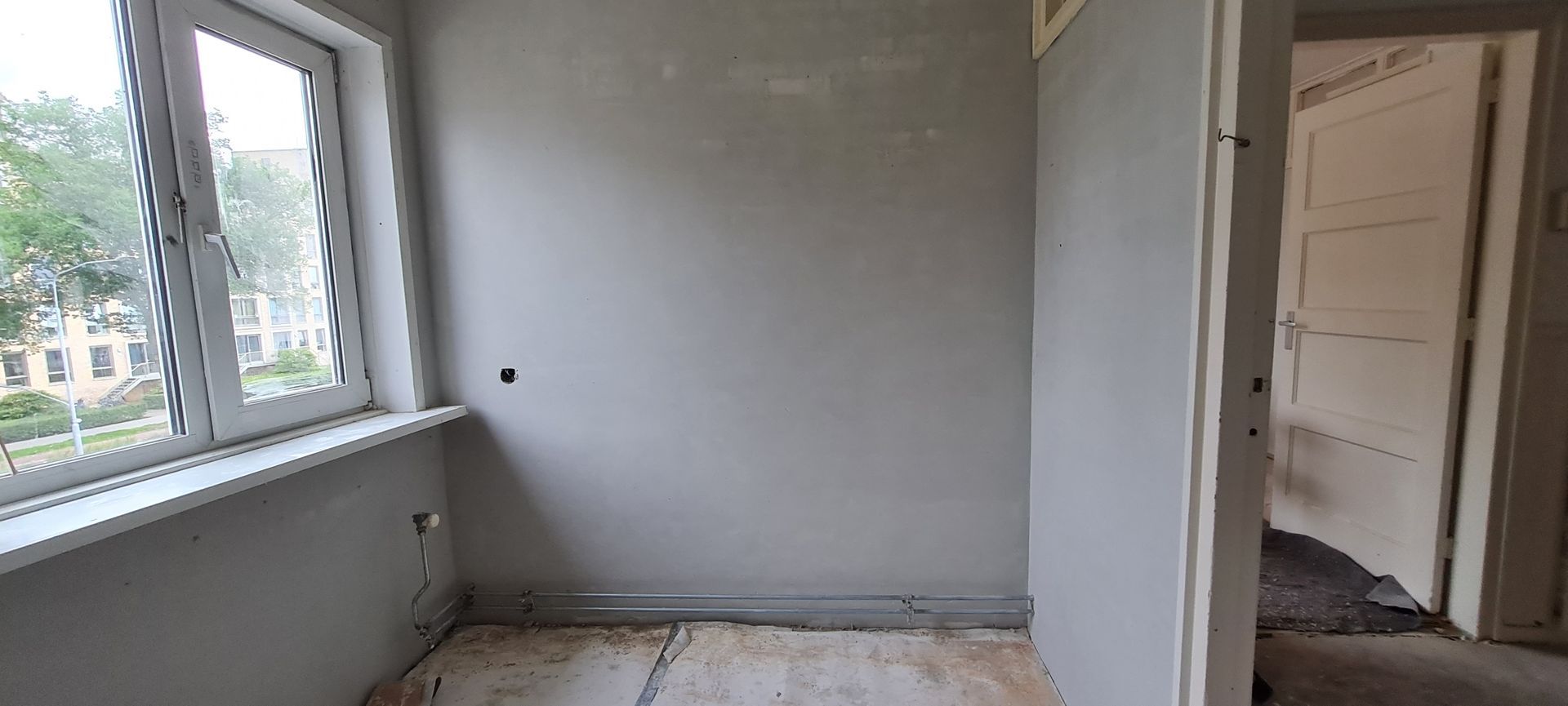 Een lege kamer in renovatie met grijze muren, een raam dat licht binnenlaat en een deur naar een andere kamer.