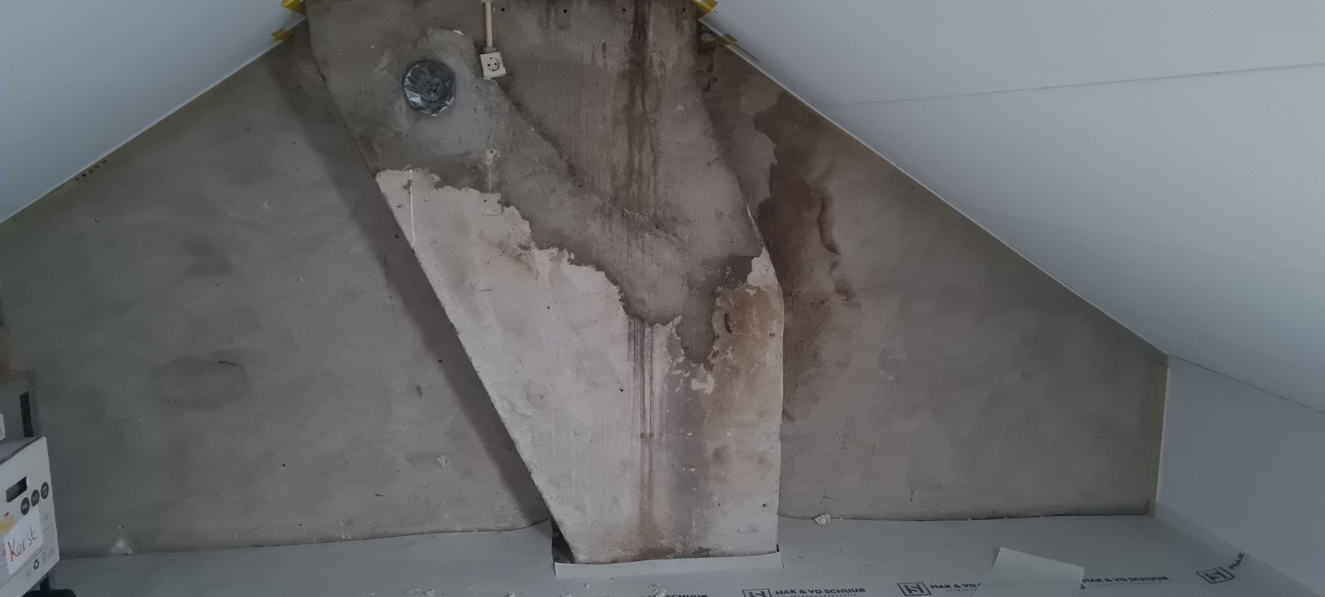 Beschadigde zolderwand met afbladderend stucwerk en een zichtbare bruine vlek onder een schuin plafond.