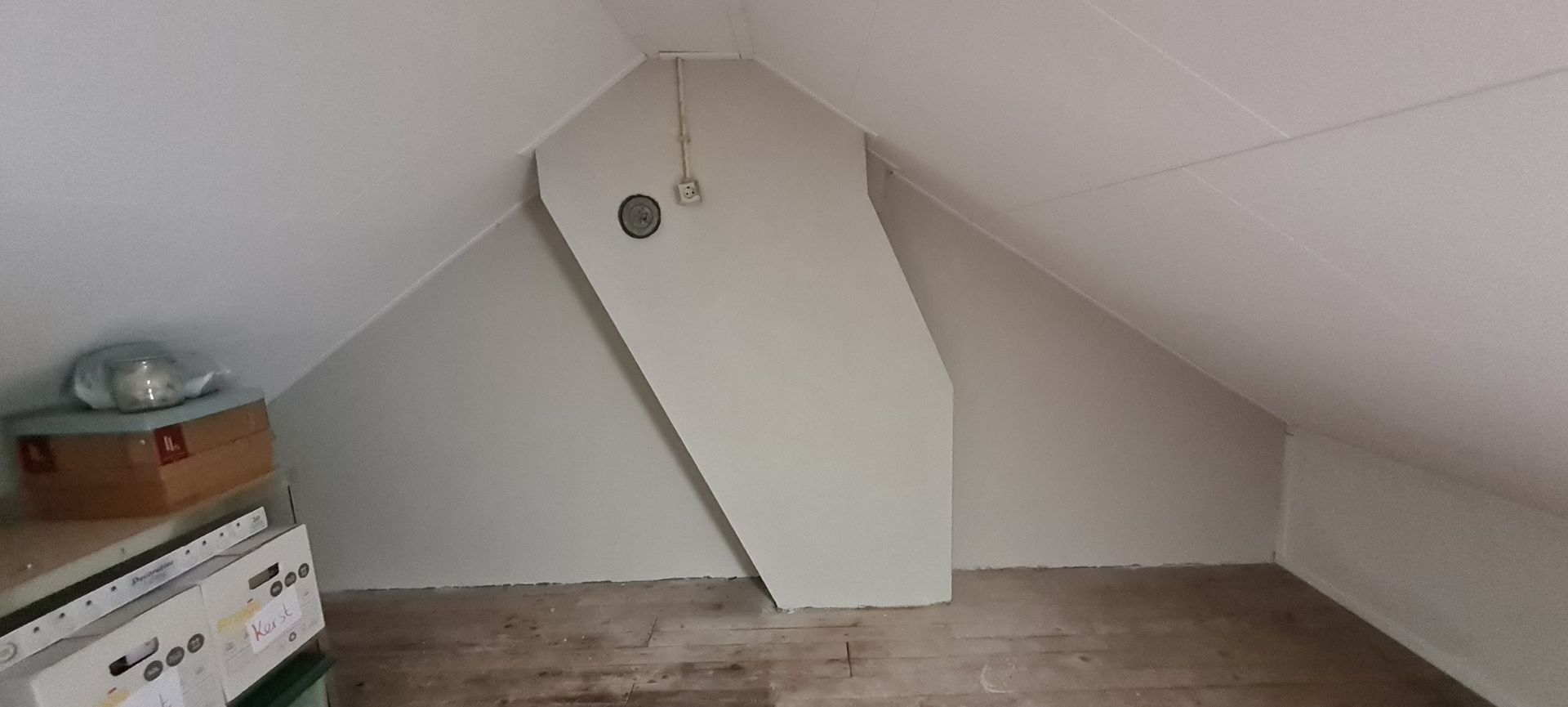 Kleine zolderkamer met een schuin wit plafond, houten vloer en opbergdozen langs de linkerwand.