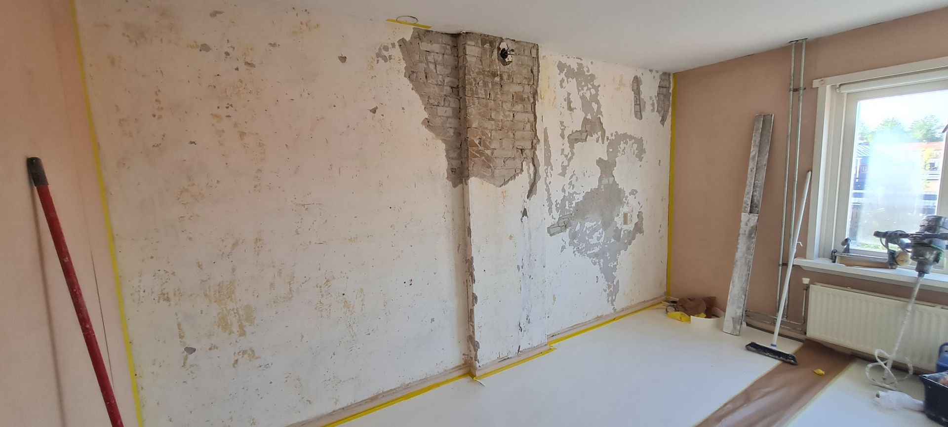 Een gedeeltelijk ontblote binnenmuur met afbladderend stucwerk, een ladder en een raam in een renovatieruimte.