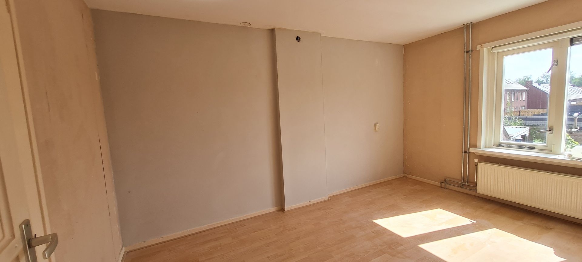 Lege beige kamer met houten vloer, zonlicht door het raam en een radiator eronder.